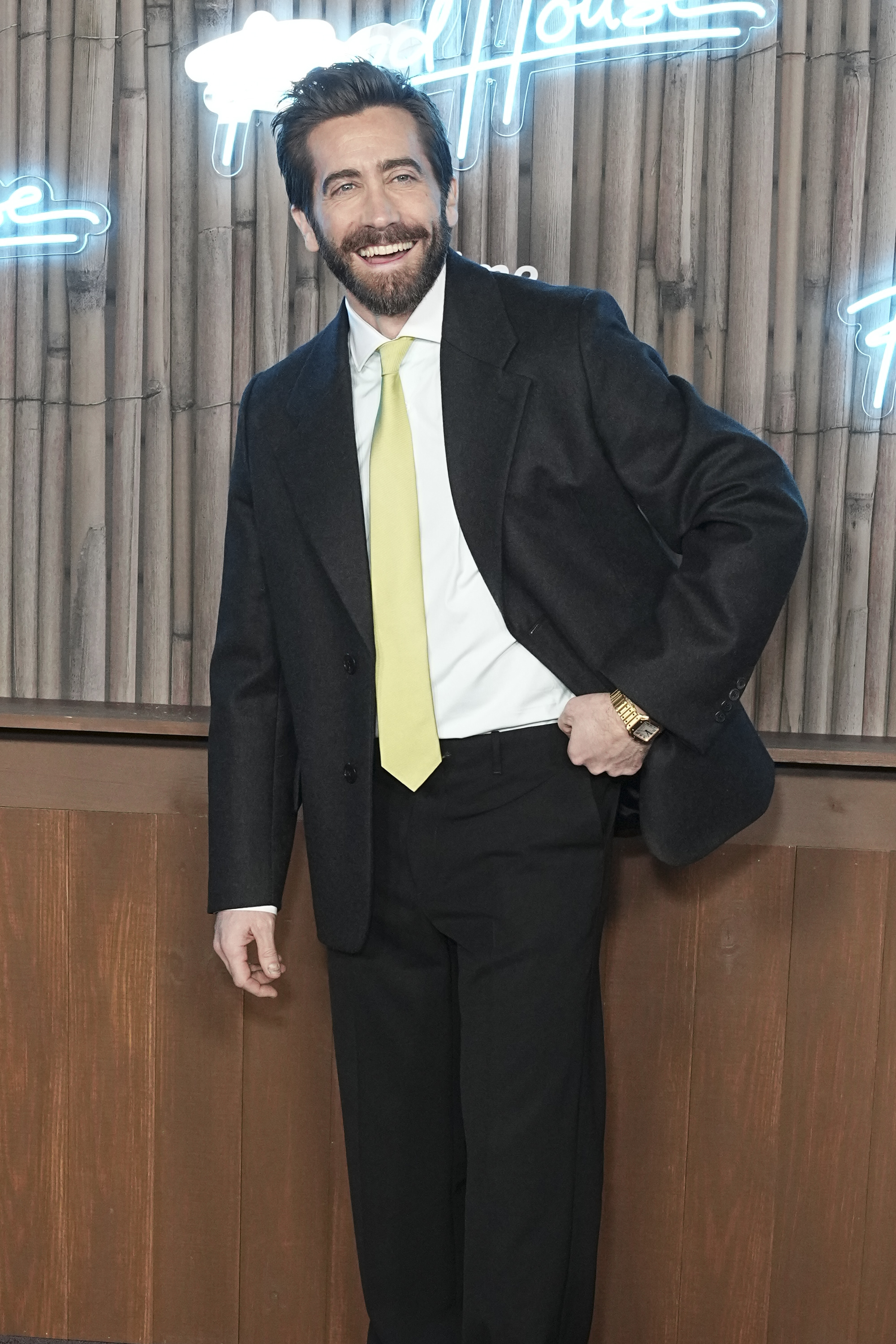 Jake Gyllenhaal smiles while wearing a dark suit with a yellow tie, standing in front of a wooden wall with neon signs