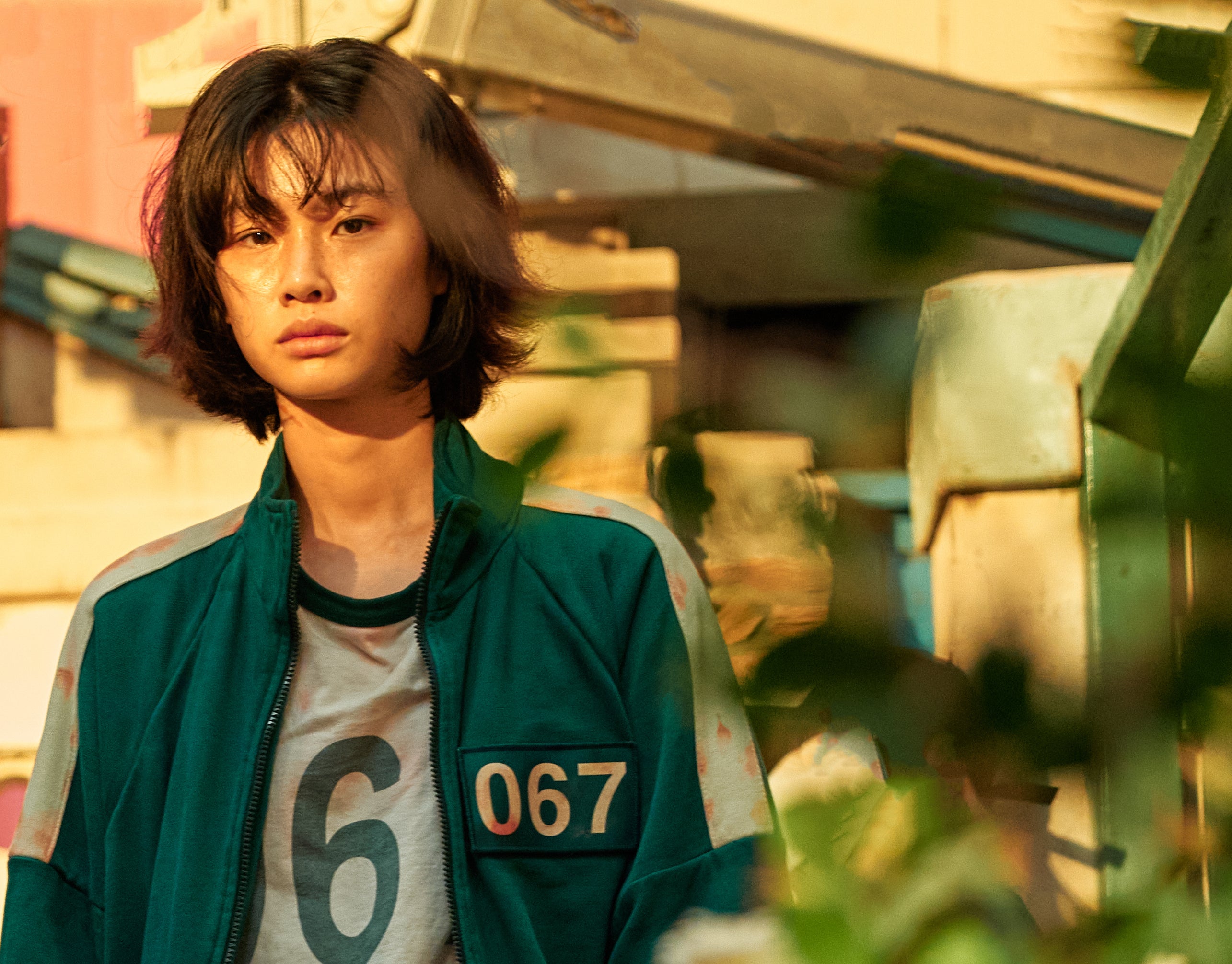 HoYeon Jung in 'Squid Game' wearing a sports jacket and t-shirt with number 067, standing outdoors surrounded by greenery