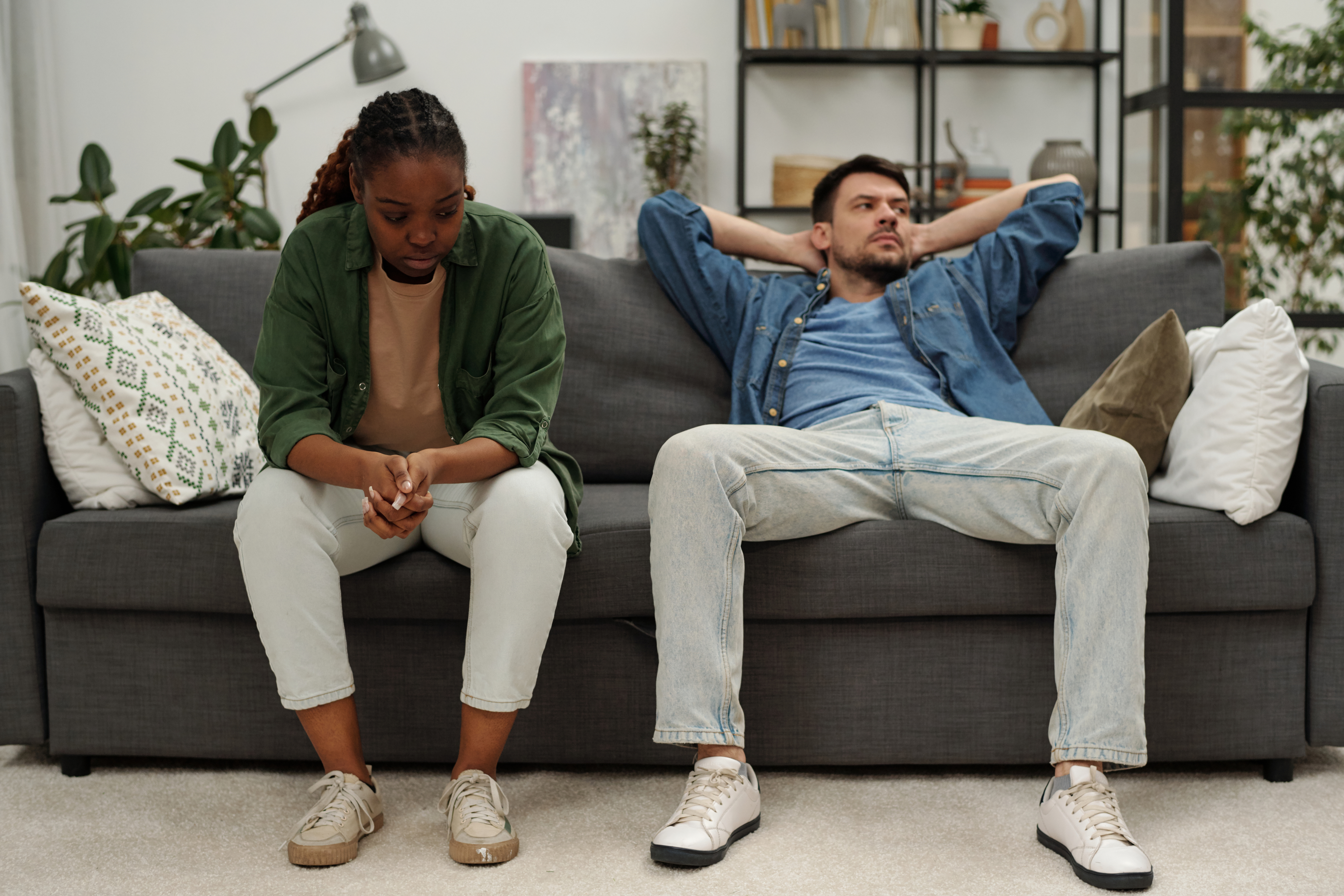 A man and woman sit on a couch; the woman looks down and clasps her hands, while the man leans back, arms crossed behind his head