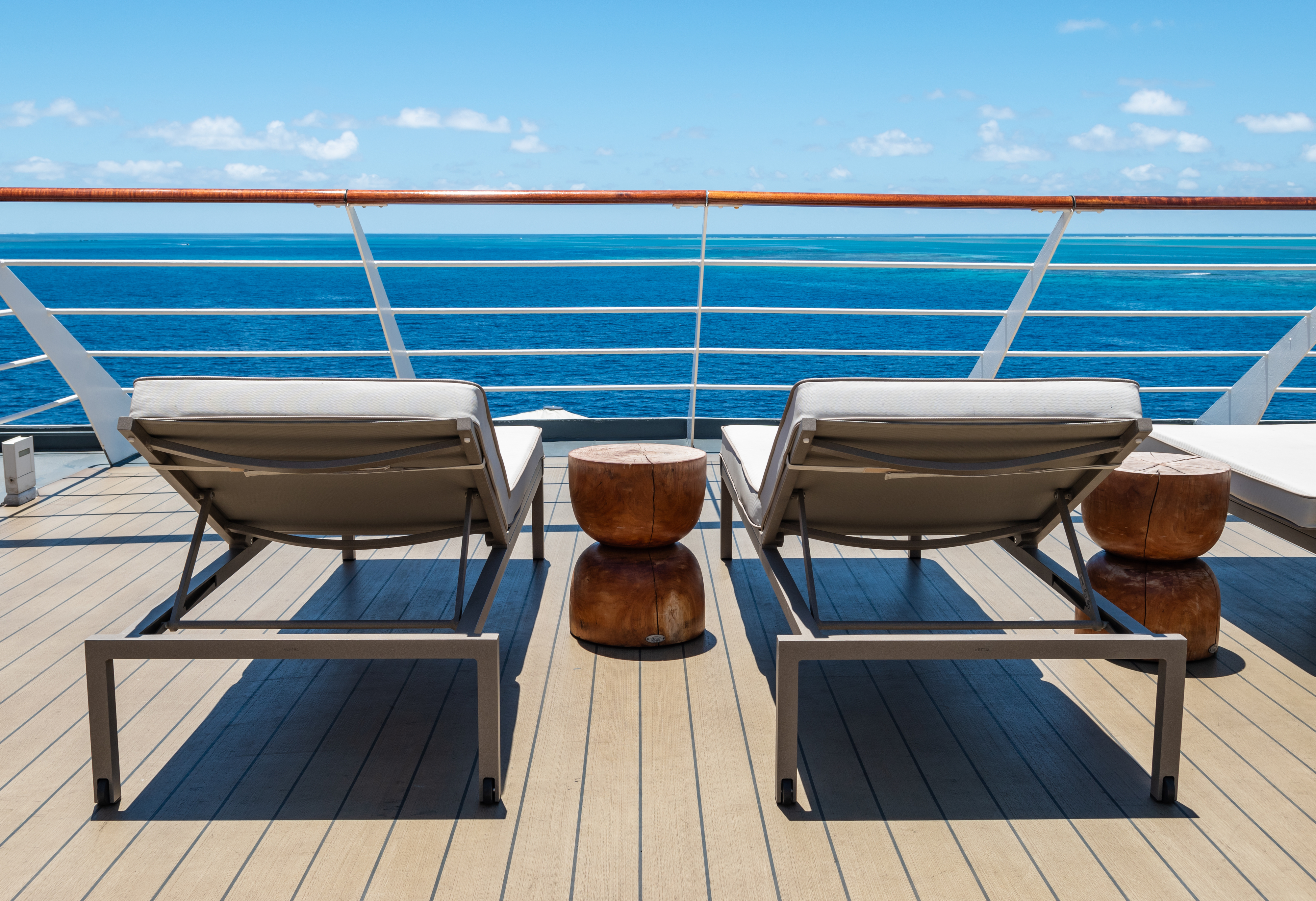 Two lounge chairs and small wooden stools facing the sea on a sunny cruise ship deck. Blue ocean stretches out to the horizon. No people are present