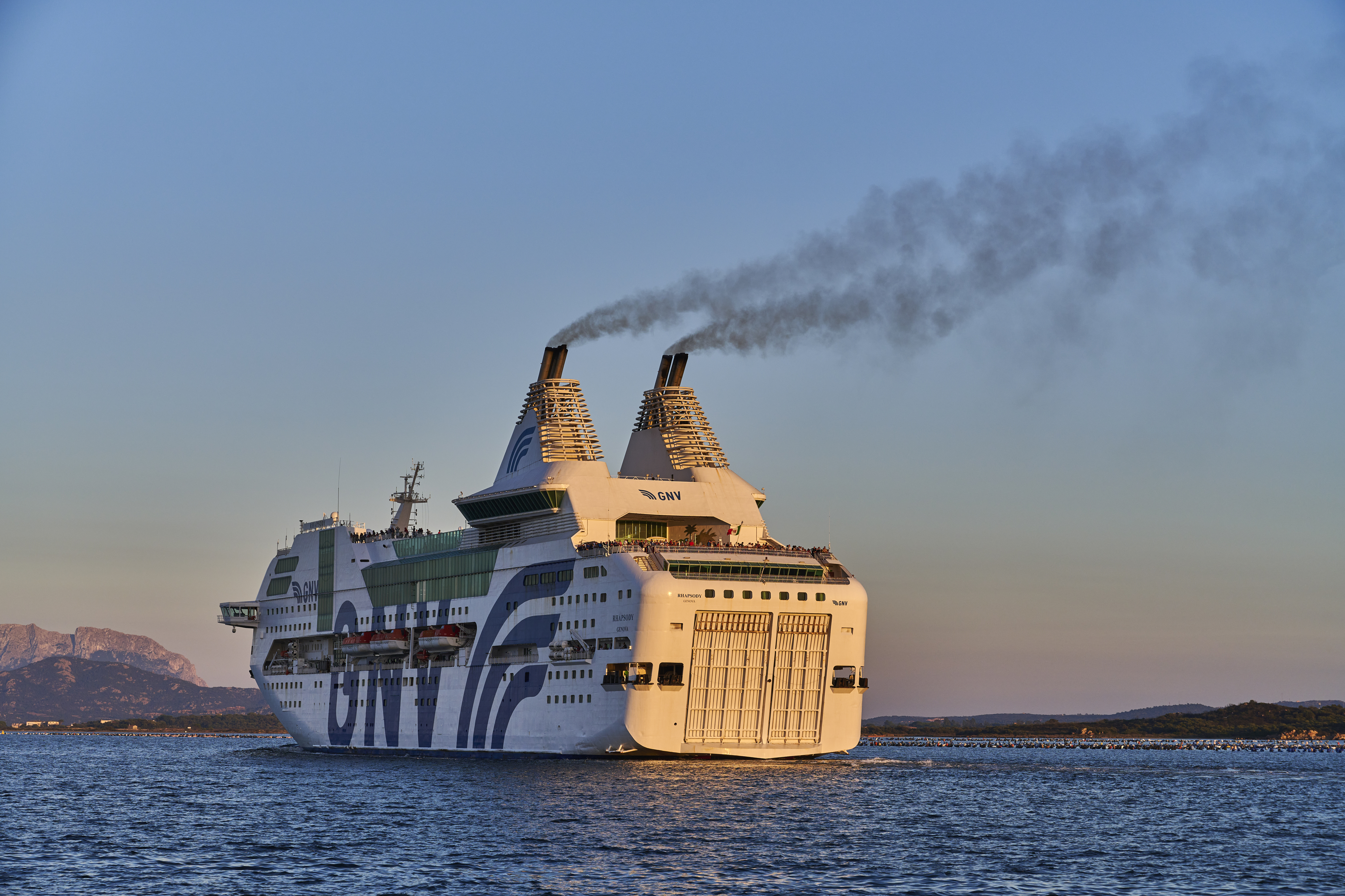 A large cruise ship with the name &quot;GNV&quot; is sailing through calm waters during what appears to be either sunrise or sunset, with mountains visible in the background