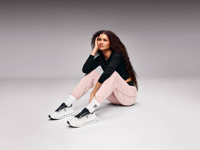 Zendaya sitting on the floor, wearing a black long-sleeve top, light-colored pants, and white sneakers, looking off to the side