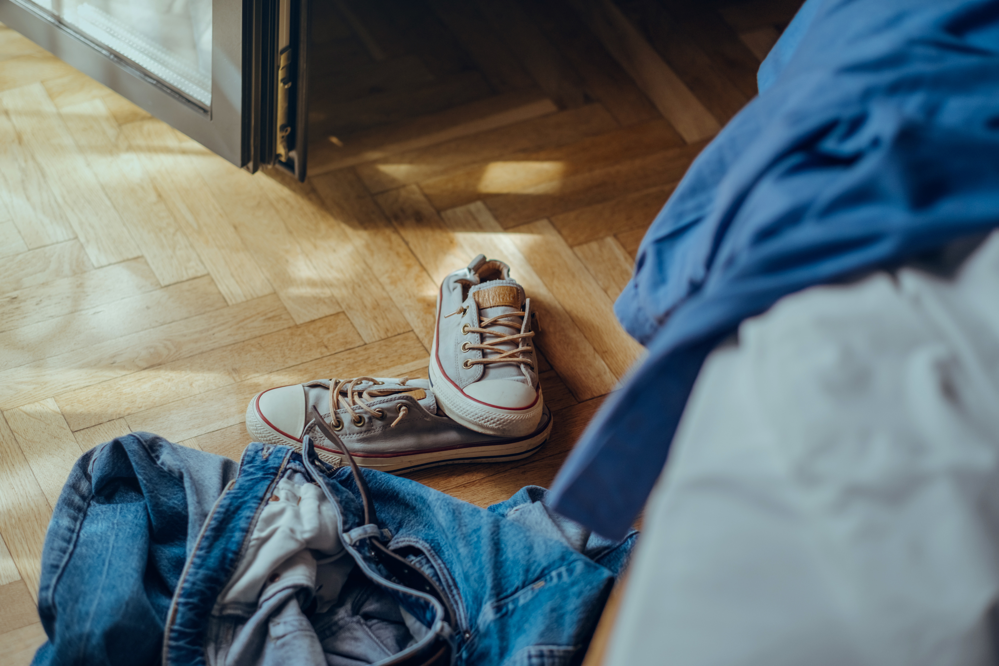 A pair of sneakers placed on a wooden floor next to scattered clothing, including jeans and a blue shirt, near an open door