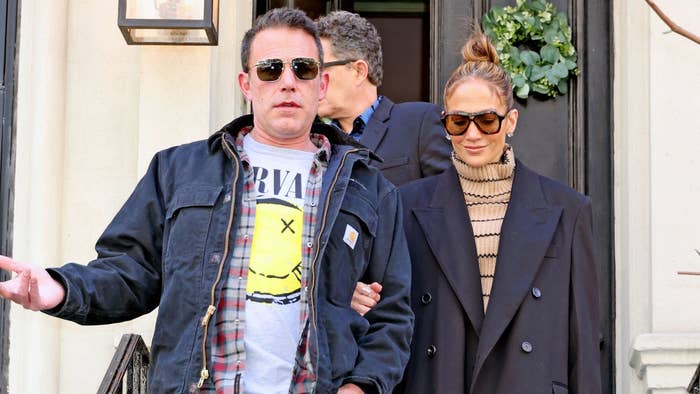 Ben Affleck in a casual outfit with a Nirvana tee, flannel, and jacket, and Jennifer Lopez in a chic coat and glasses, both outside a building
