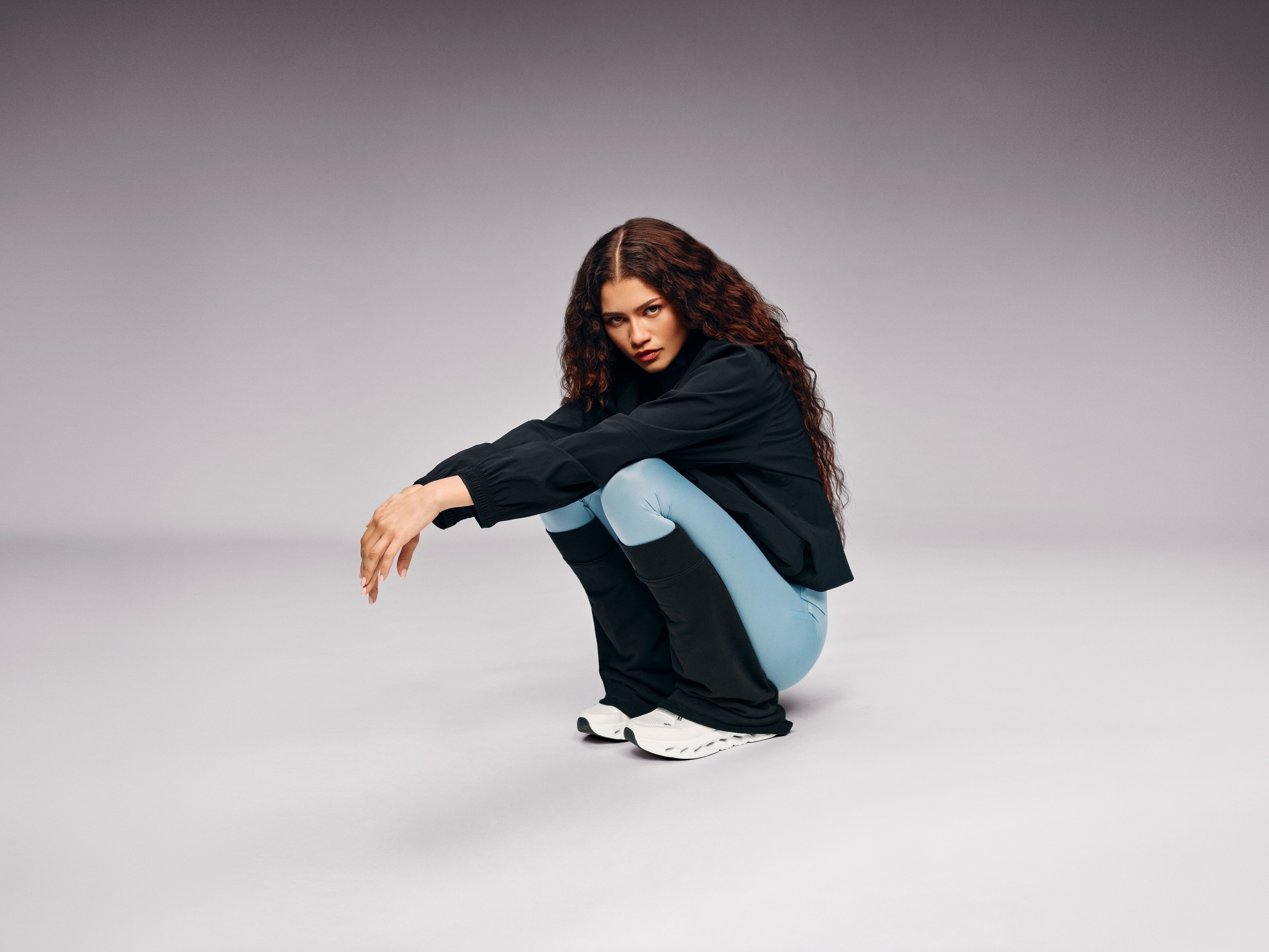 Zendaya poses in stylish activewear, pairing a black top with light blue leggings and white sneakers, set against a minimalist backdrop