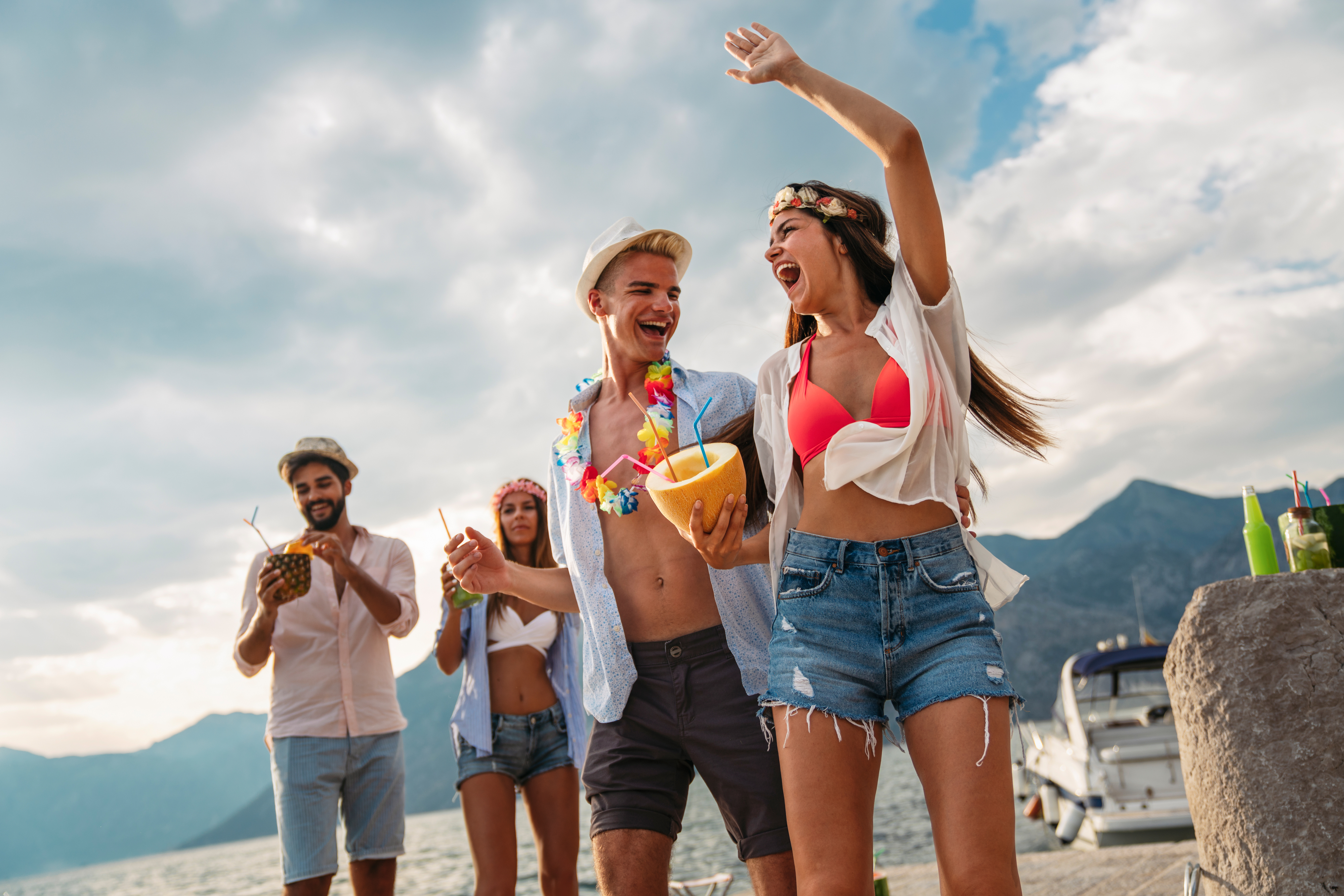Group of friends enjoying a tropical vacation by the beach, wearing summer clothes, holding drinks, and dancing with a boat and mountains in the background