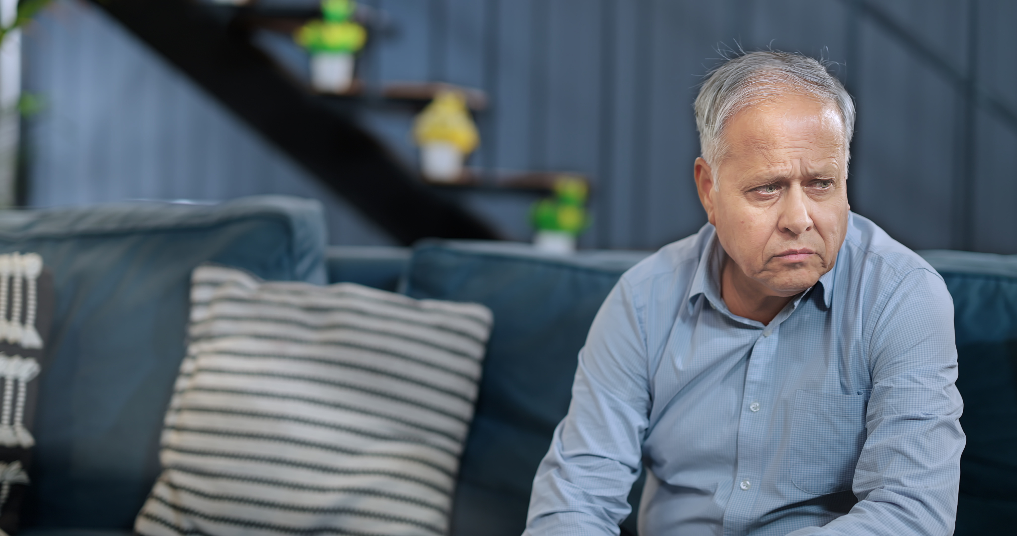 Older man with grey hair sits on a sofa looking pensive, wearing a light shirt in a modern living room