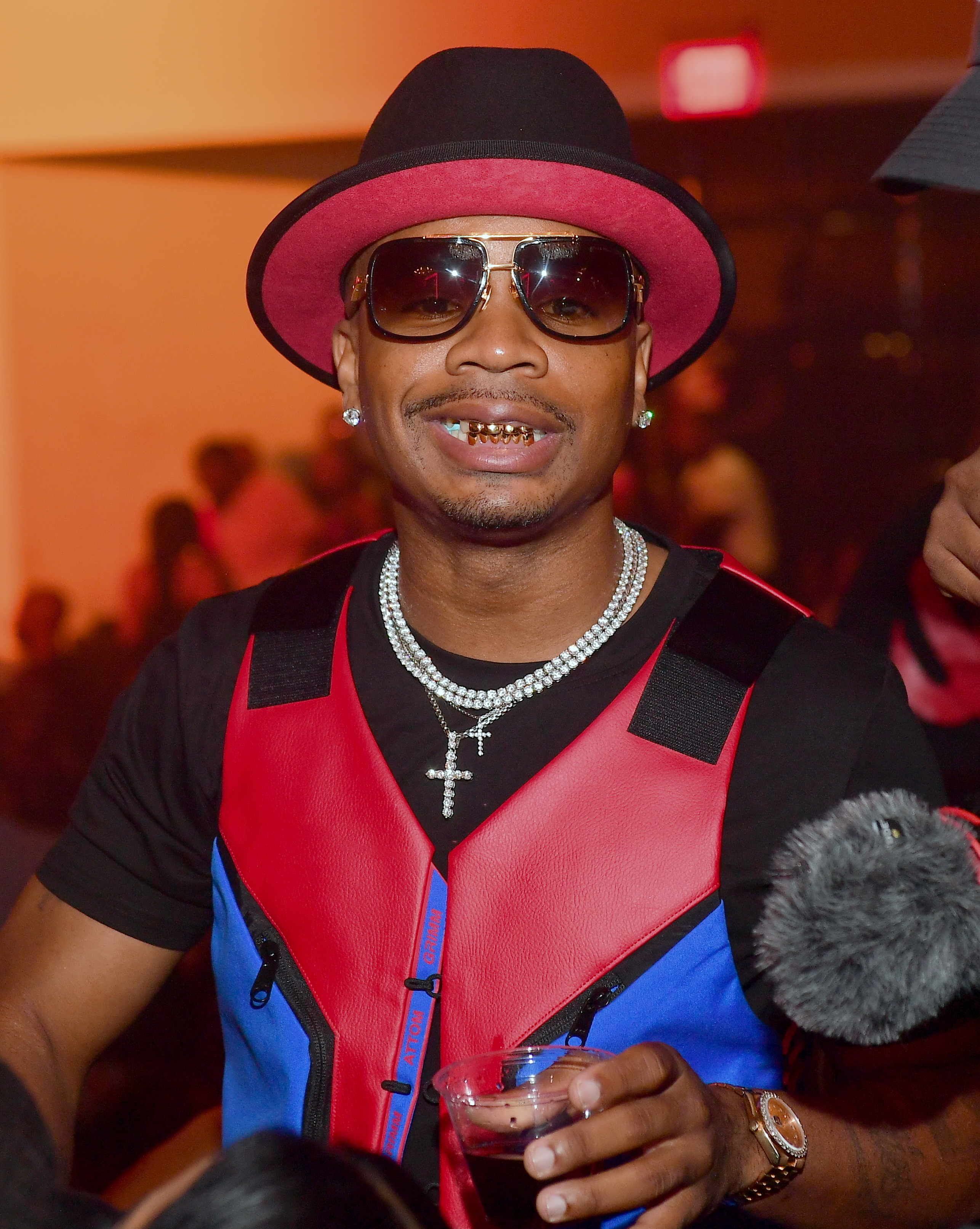 Plies wearing sunglasses, a red and black hat, a red and blue vest, and a diamond necklace, holds a drink at an event