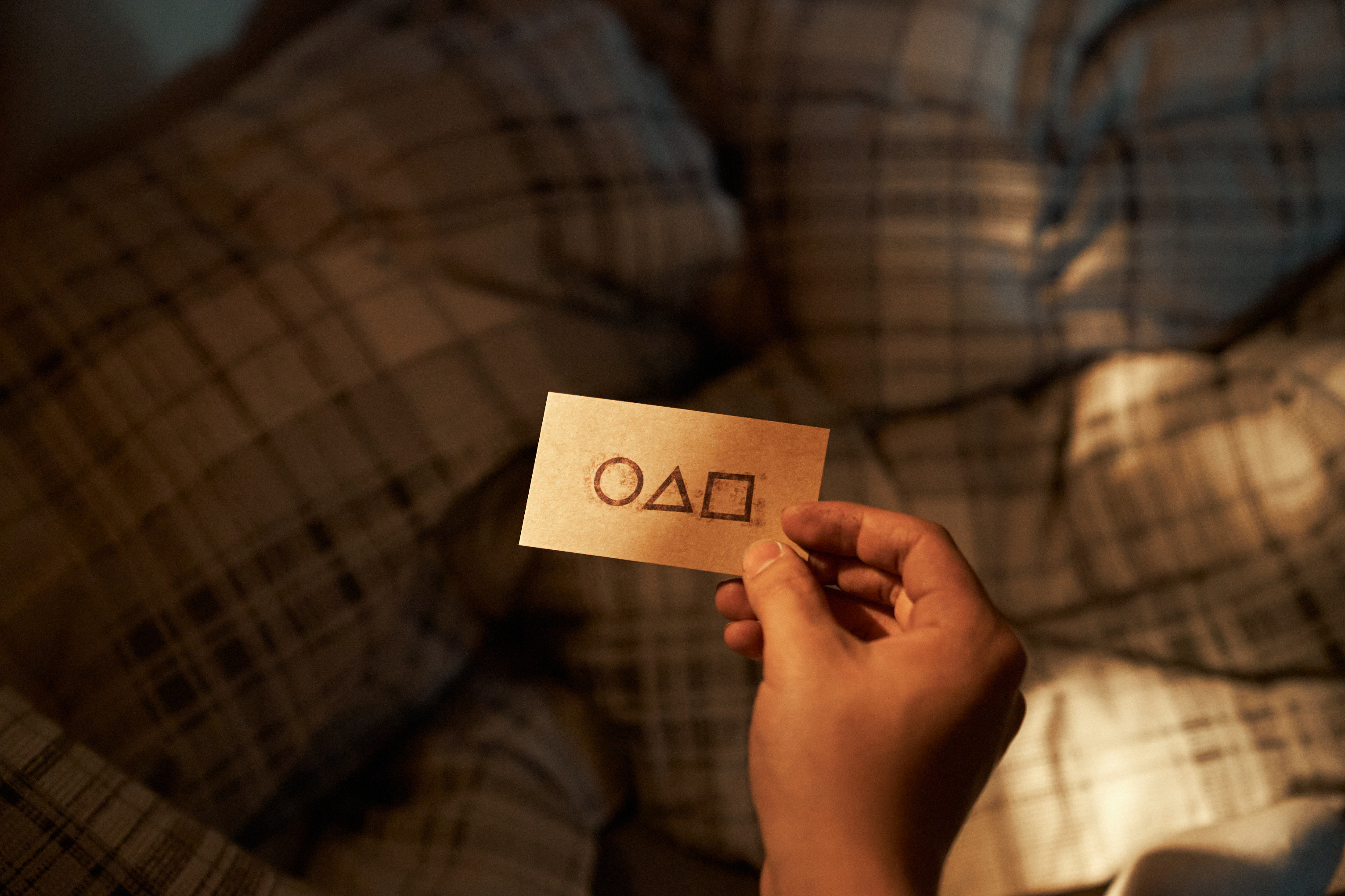 A hand holds a card with symbols of a circle, triangle, and square, resting on a plaid bedspread