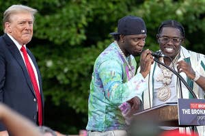 Donald Trump stands next to rappers Fresha and Symphony at a podium during an outdoor event. Fresha and Symphony are speaking into microphones
