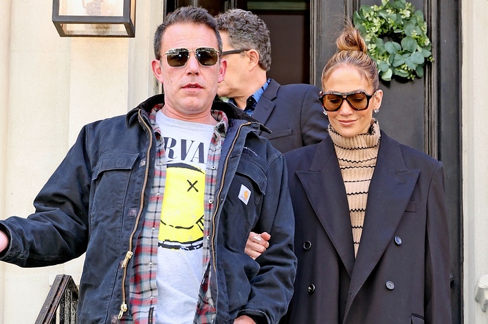 Ben Affleck in a casual outfit with a Nirvana shirt, and Jennifer Lopez in a stylish coat and striped turtleneck, both wearing sunglasses, walking down stairs