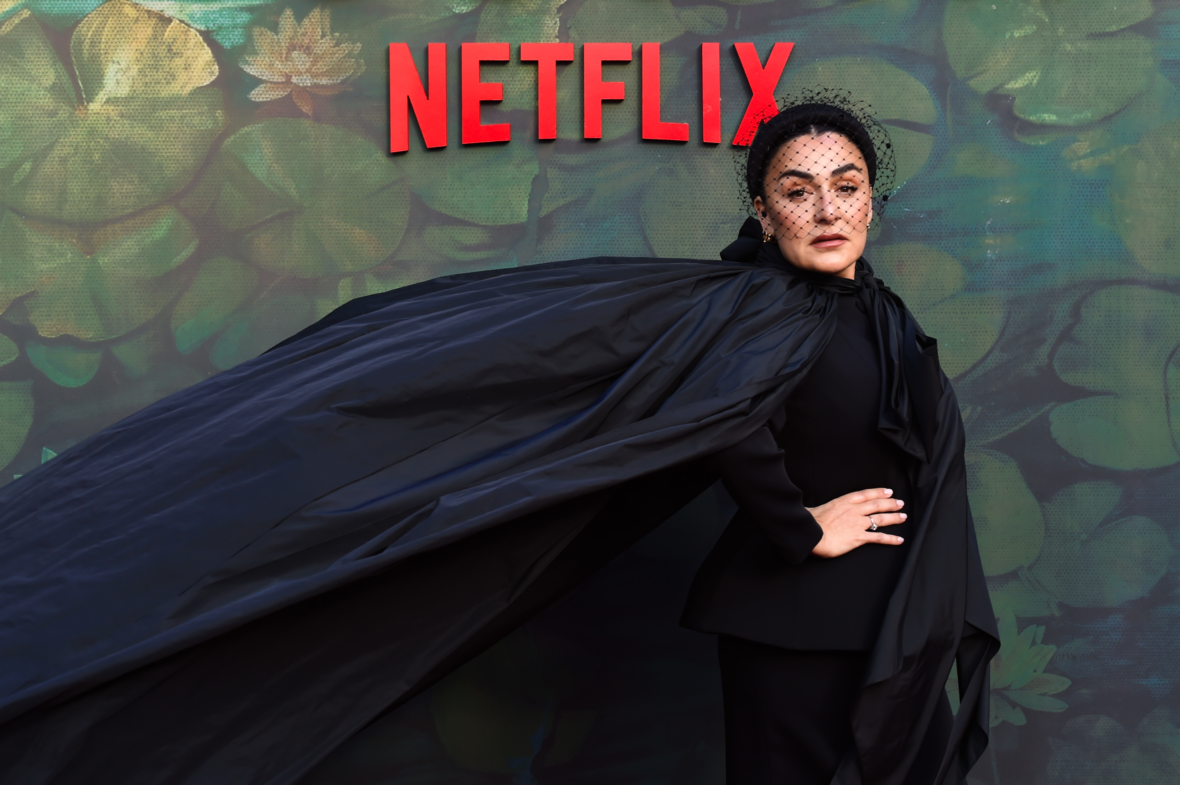 Candela Peña poses confidently on a Netflix event red carpet, wearing an elegant, flowing black outfit with a headpiece featuring a netted veil on the red carpet premiere of &quot;The Asunta Case.&quot;