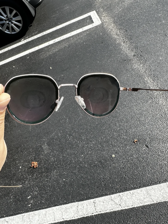 A hand holds a pair of round-framed sunglasses above a parking lot