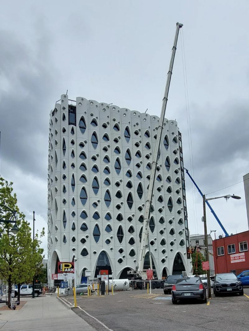 A tall, uniquely-designed building with numerous arch-shaped windows is under construction, with cranes and construction equipment surrounding it on a city street