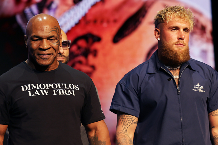 Mike Tyson (left) in a "Dimopoulos Law Firm" t-shirt and Jake Paul (right) in a dark zip-up jacket, posing at an event