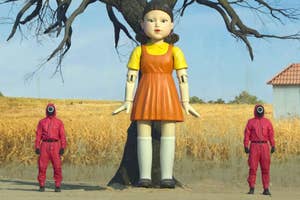 The image shows a large doll from Squid Game standing near a tree with two masked guards in red jumpsuits on either side