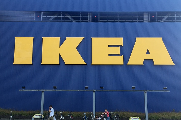 青い建物の大きな「IKEA」看板の前で、自転車が並んでいる。手前には歩いている人が二人いる。