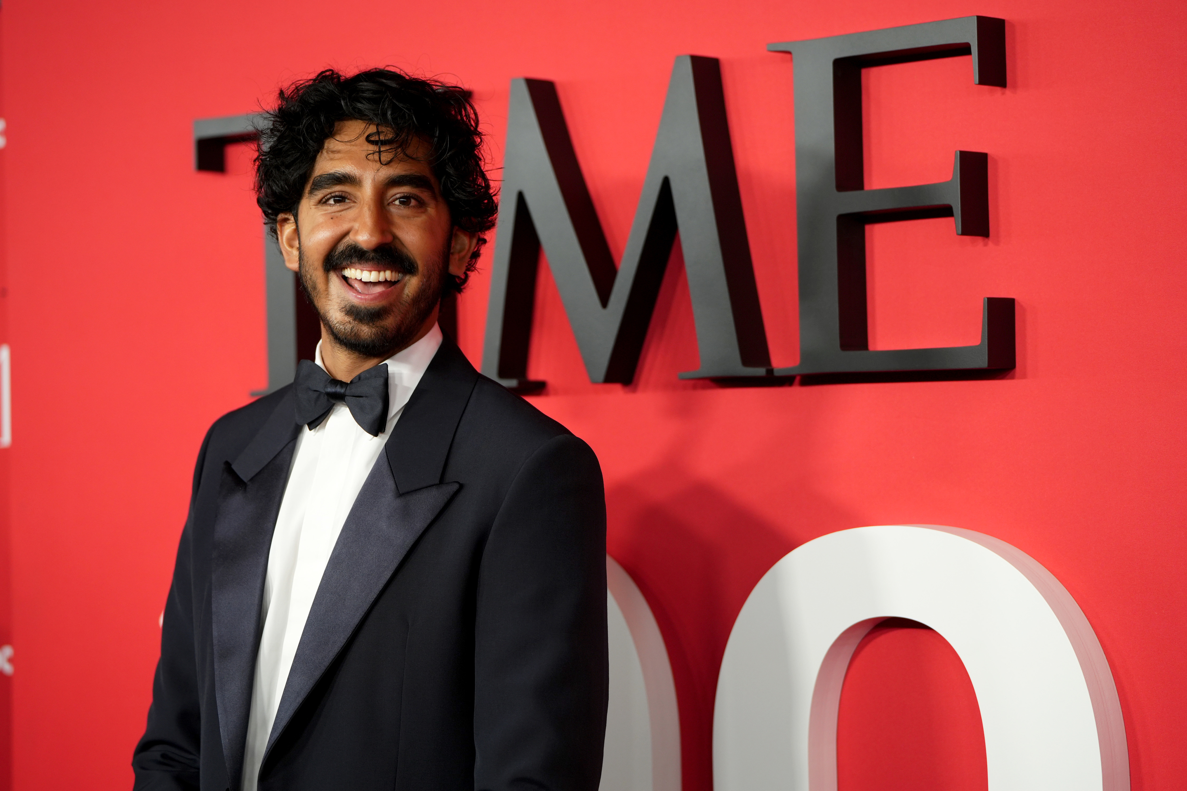 Dev Patel in a black tuxedo smiles on the red carpet by the TIME logo