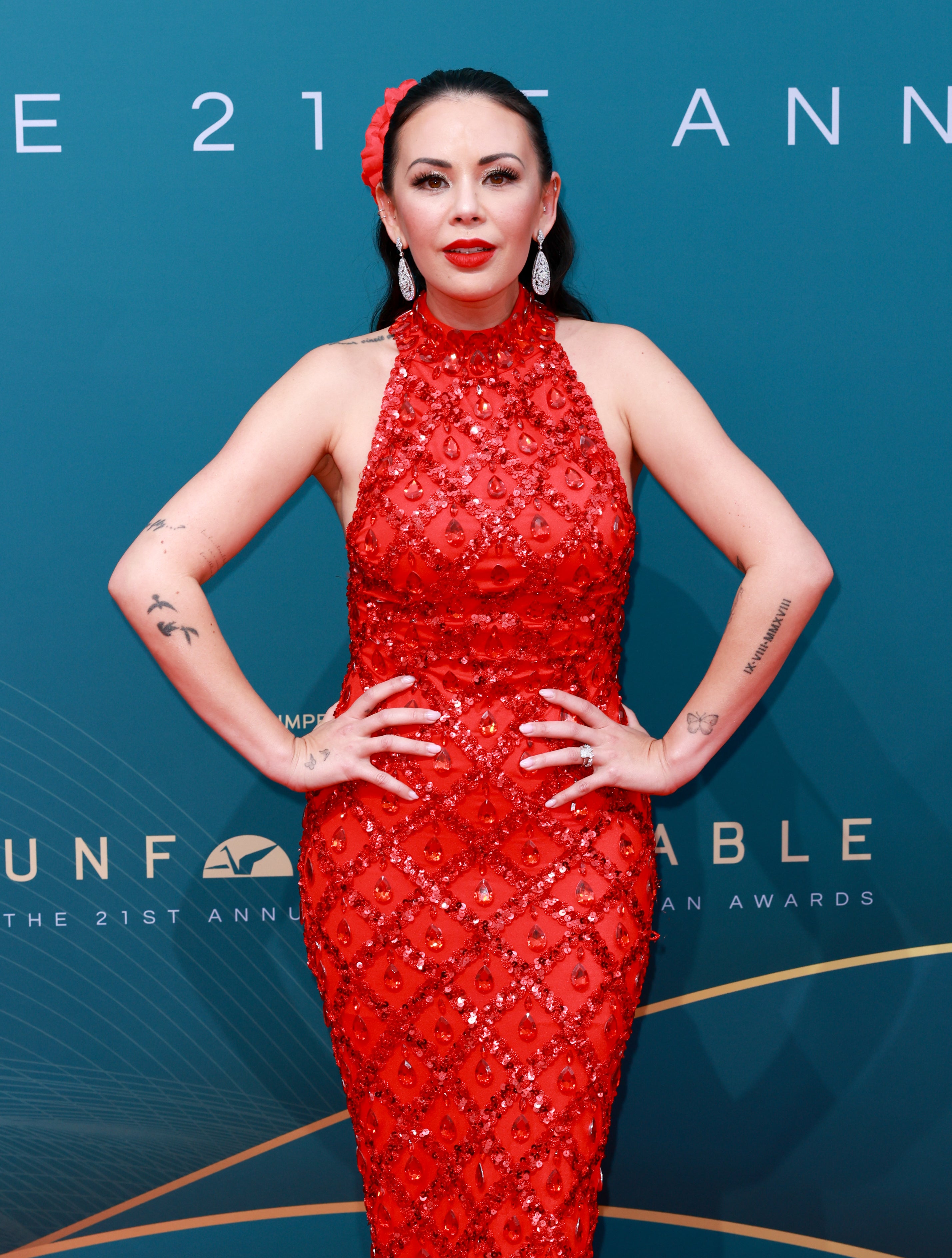 Janel Parrish in an embellished red gown with hands on hips, standing against event backdrop