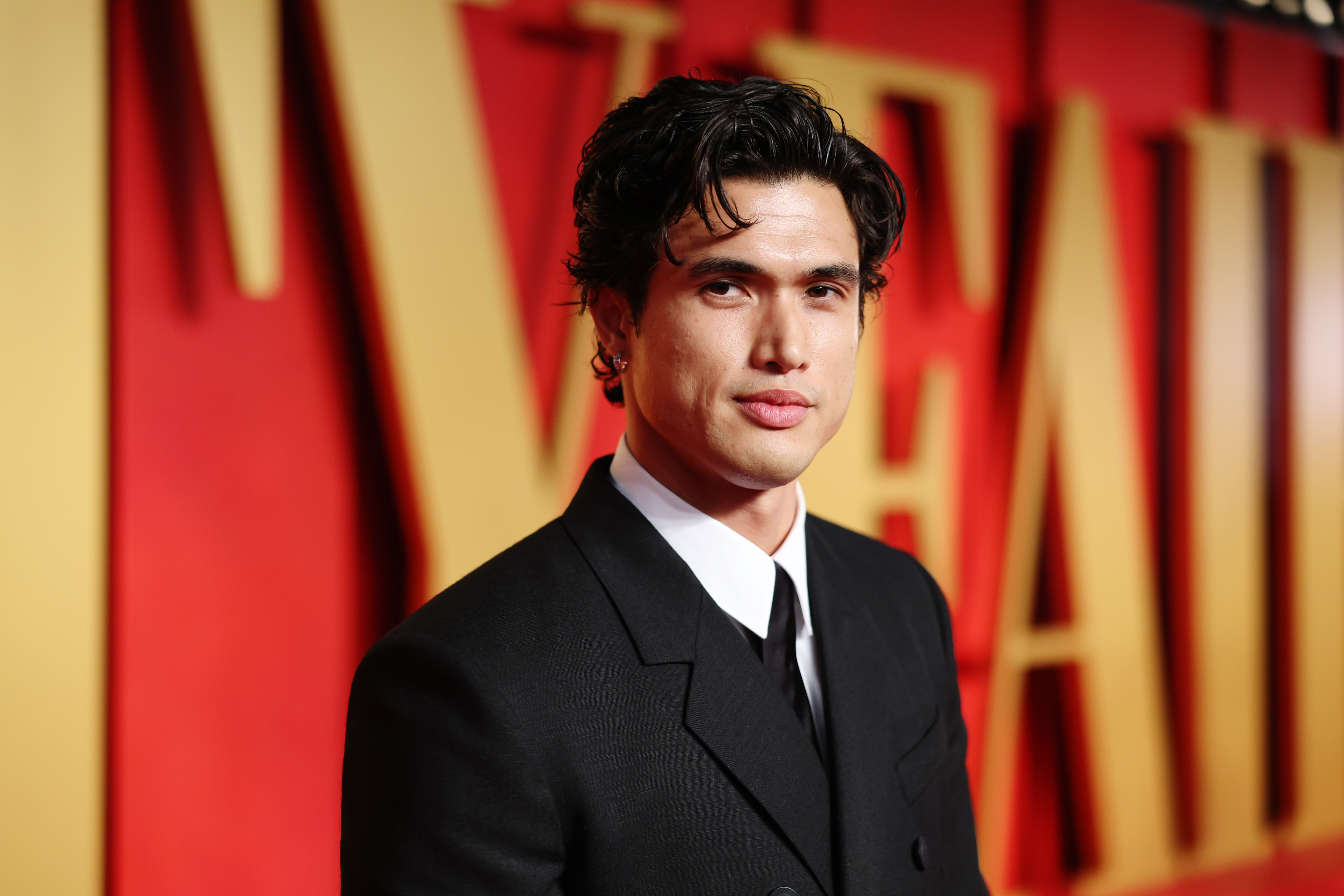 Charles Melton in a formal black suit posing before a backdrop with bold letters