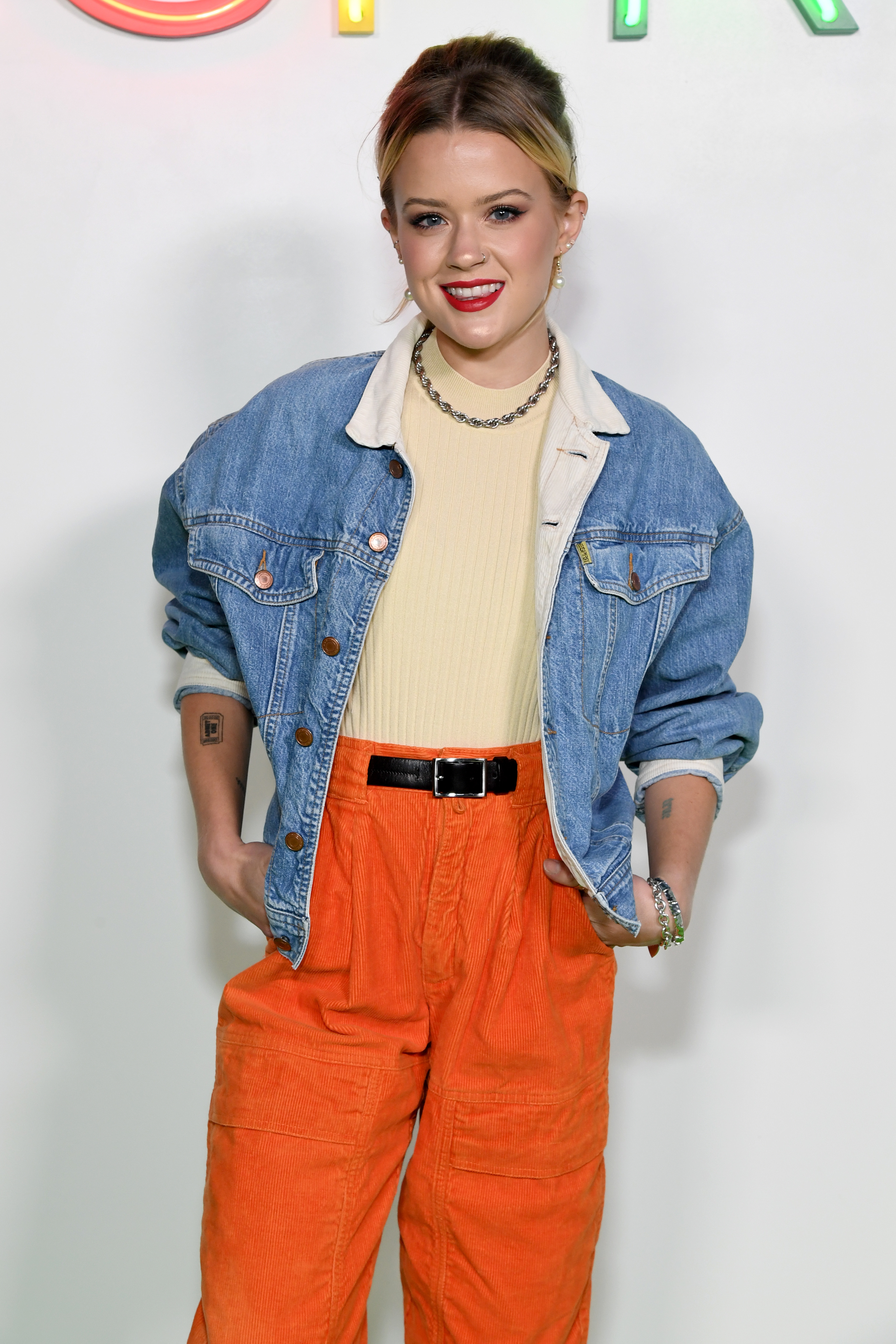 Ava Phillippe in denim jacket over a light top, orange pants, and chunky necklace, smiling