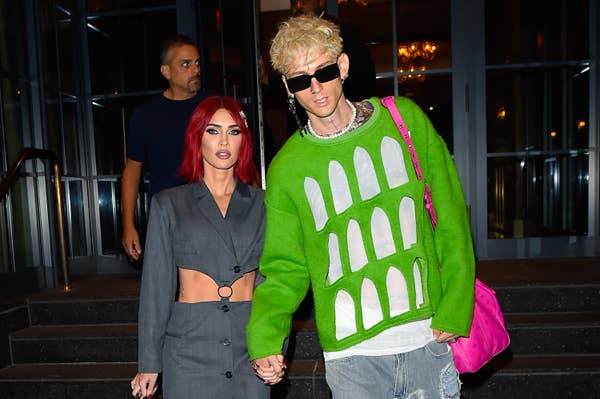 Two individuals walking, one in a green sweater with a unique pattern, the other in a cut-out blazer. They are holding hands