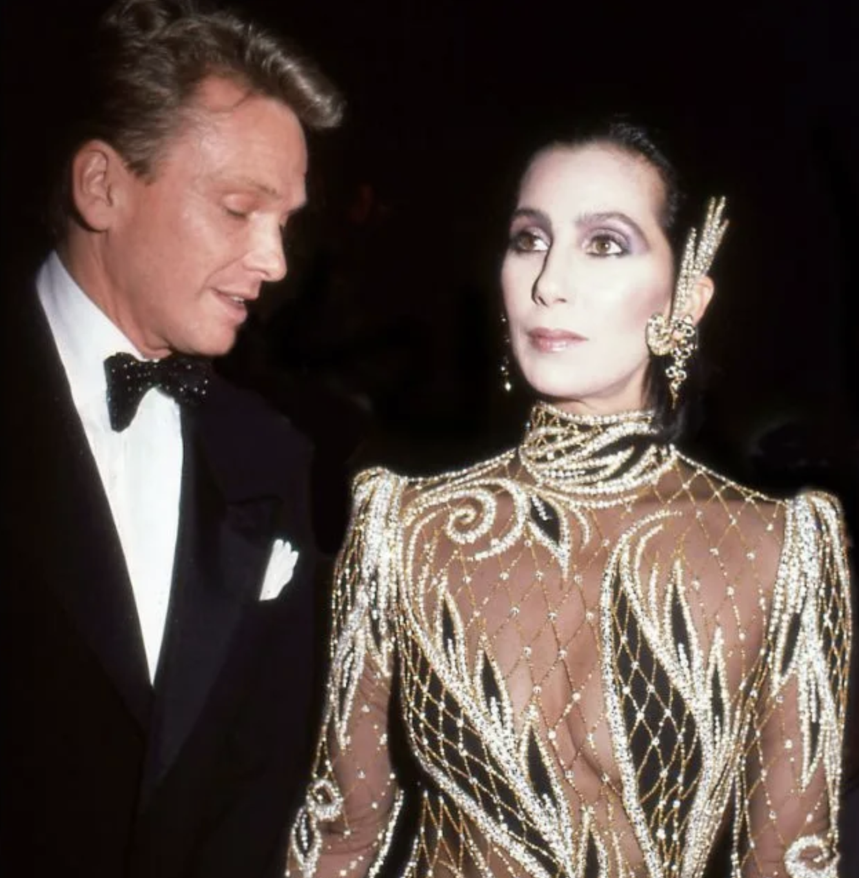 Cher con un vestido de malla y pedrería, acompañada por persona en traje formal