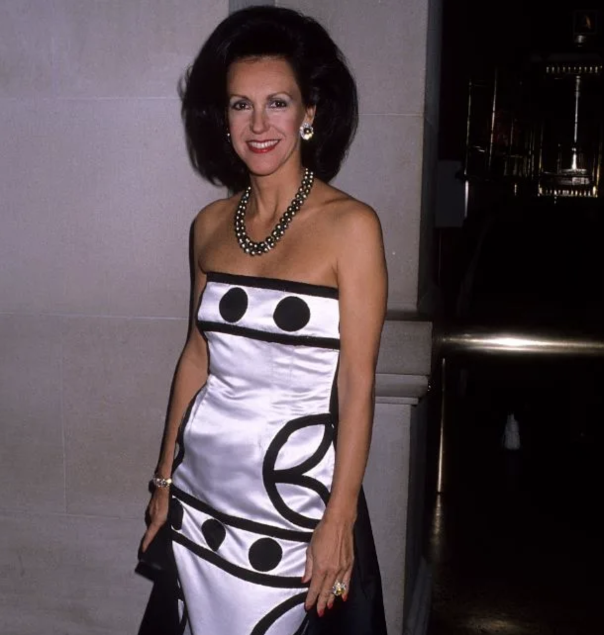 Mujer sonriendo en evento, vestido sin mangas blanco con adornos negros y collar de perlas