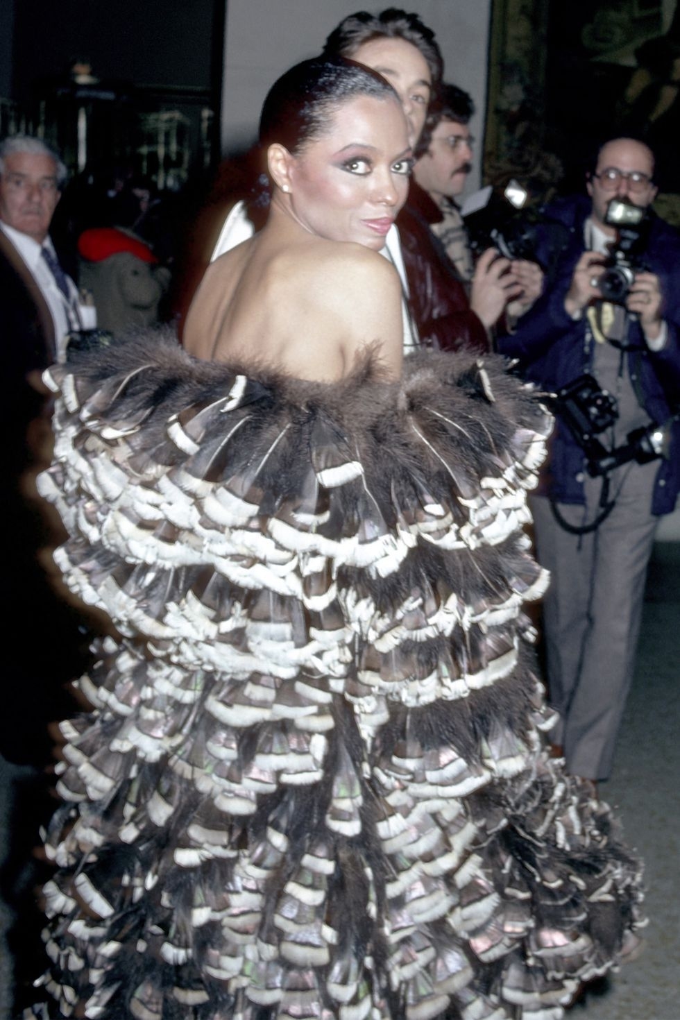 Mujer famosa en evento posando en vestido extravagante con plumas y lazos, rodeada de fotógrafos en el fondo