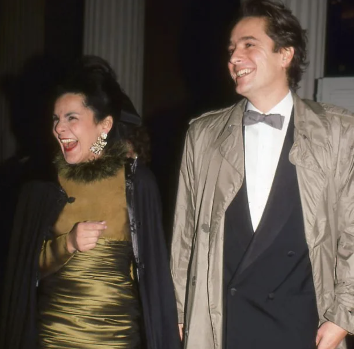 Dos personas sonriendo en evento, una mujer con vestido dorado y chaqueta negra, y hombre en esmoquin con pajarita