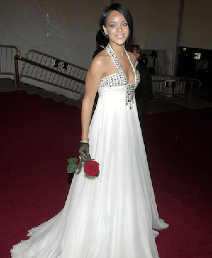 Rihanna posa con un vestido largo blanco y detalles brillantes, guante y una rosa roja en la mano