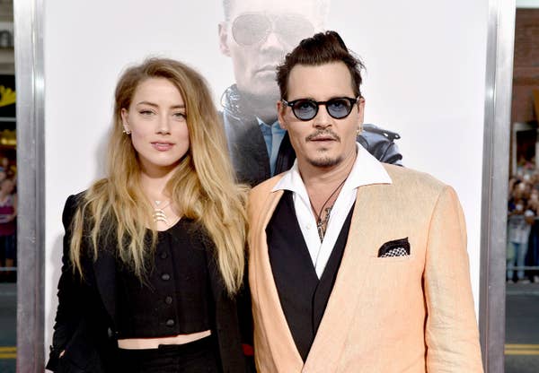 Two people posing on the red carpet; person on right in a tan suit with sunglasses, person on left in a black outfit