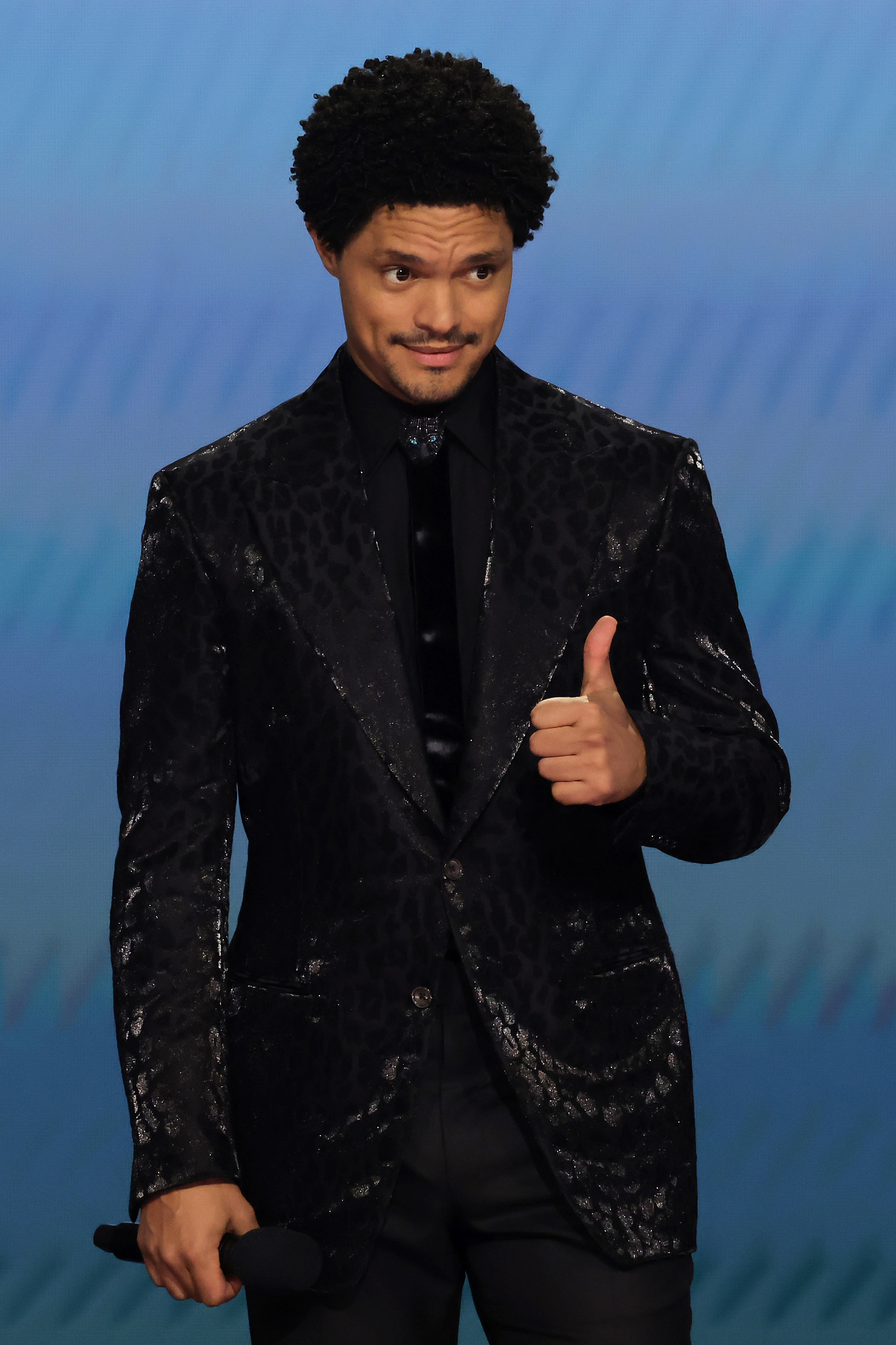 Trevor Noah in a textured blazer giving a thumbs-up onstage