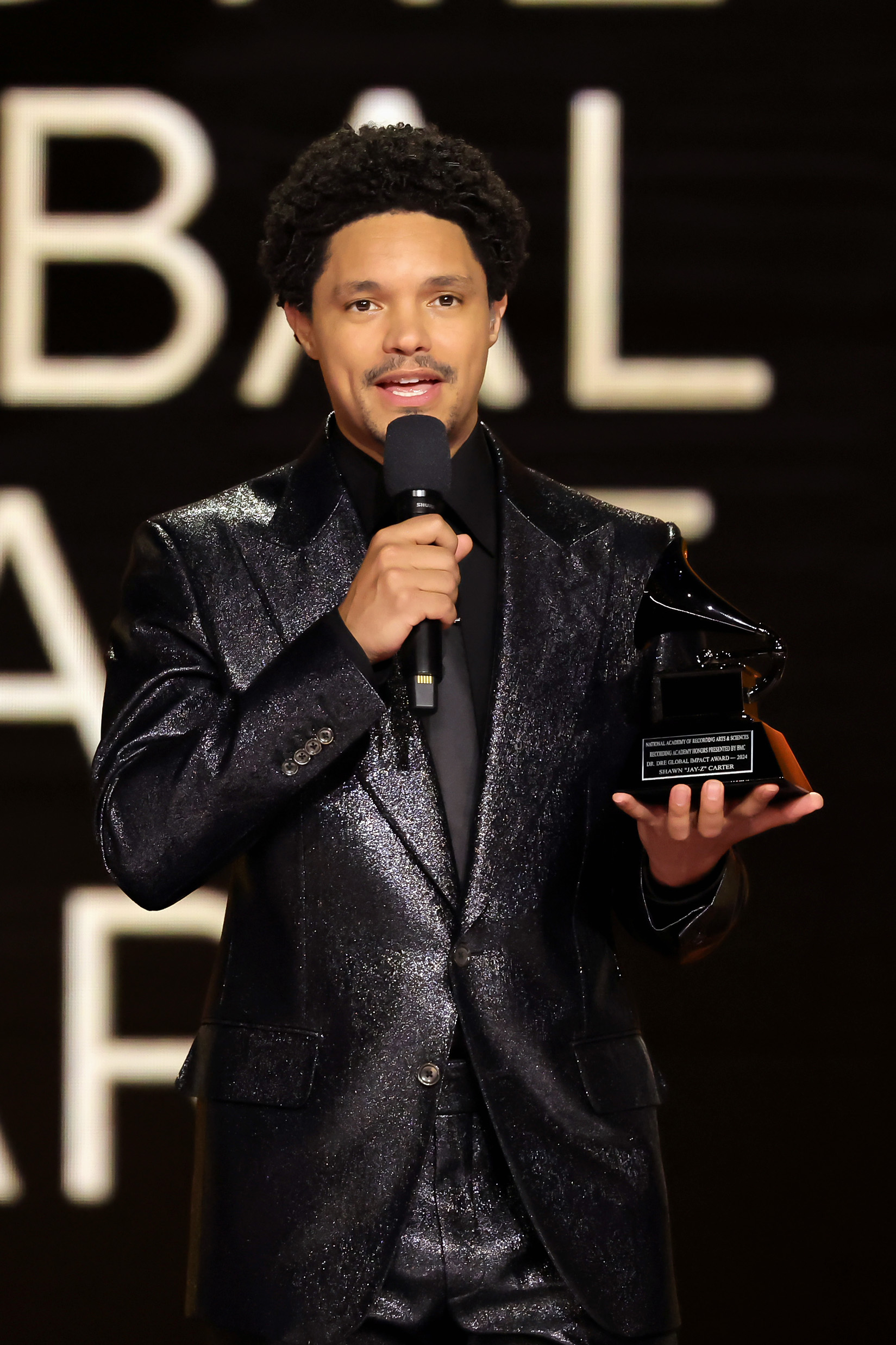 Trevor Noah in a glittery jacket holding a microphone and award on stage