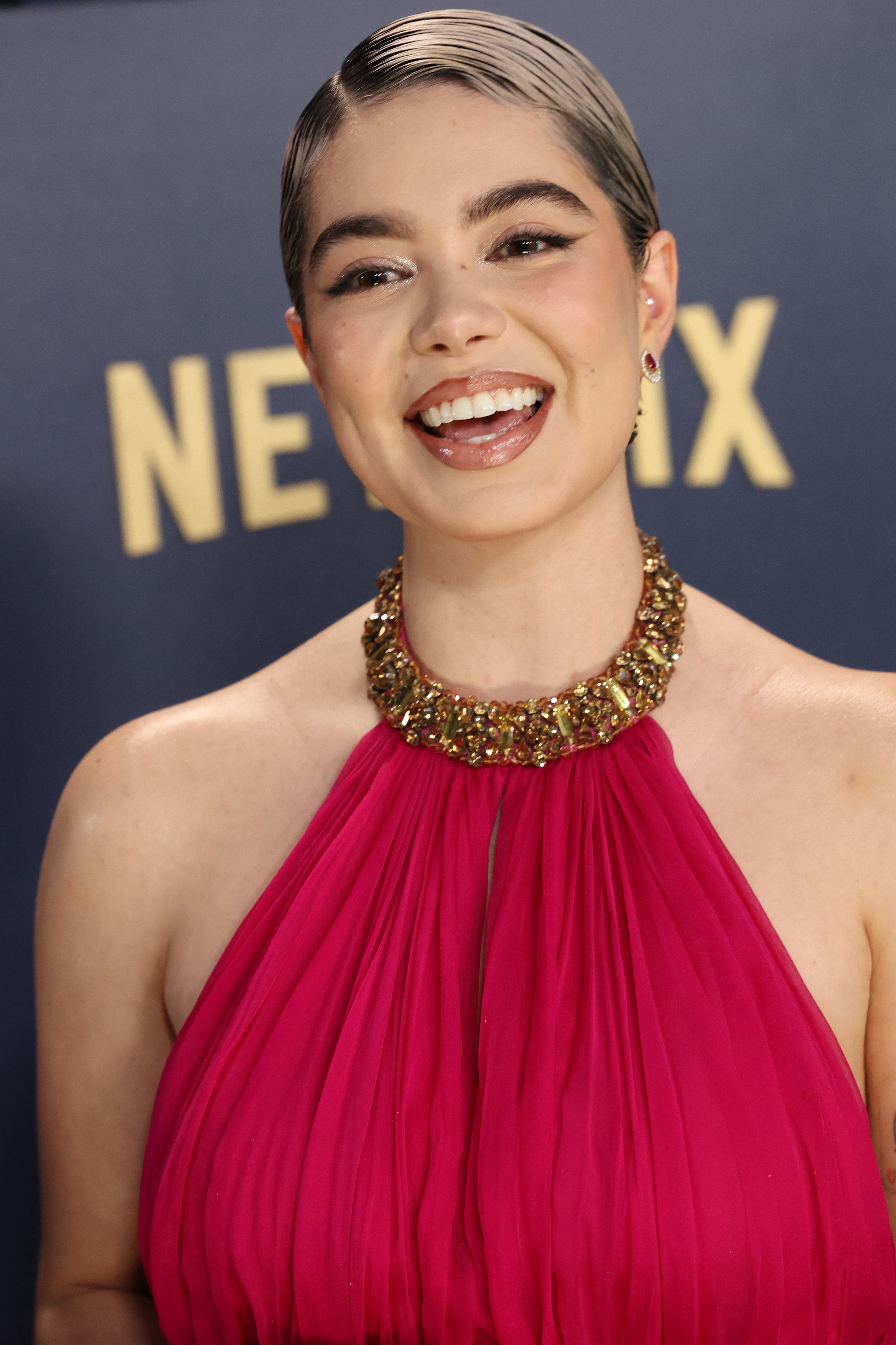 Auli&#x27;i Cravalho in pleated gown with gold neckpiece smiling against event backdrop