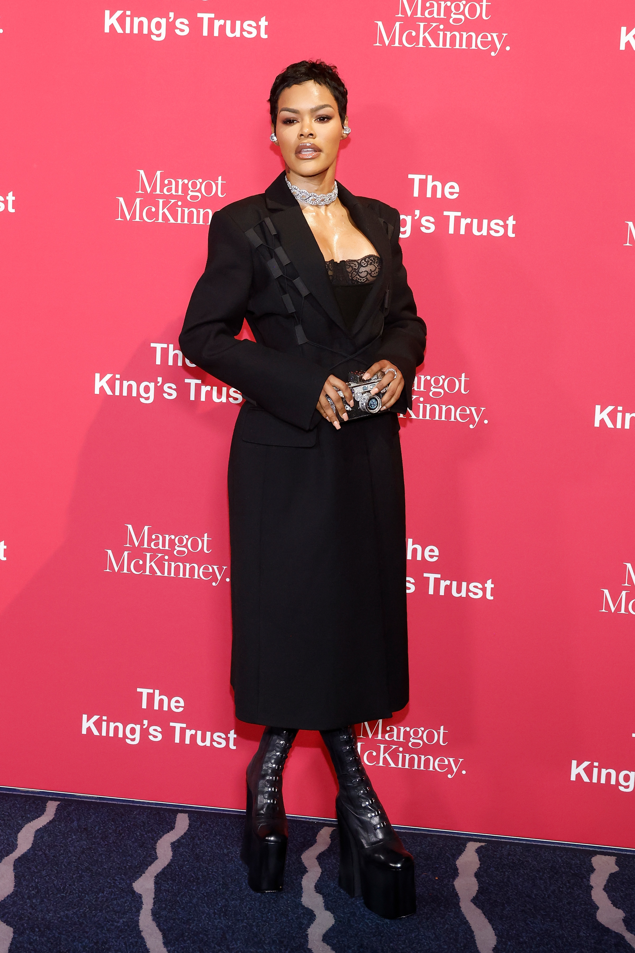 Teyana Taylor standing on red carpet in a black dress with blazer, chunky boots, and statement jewelry