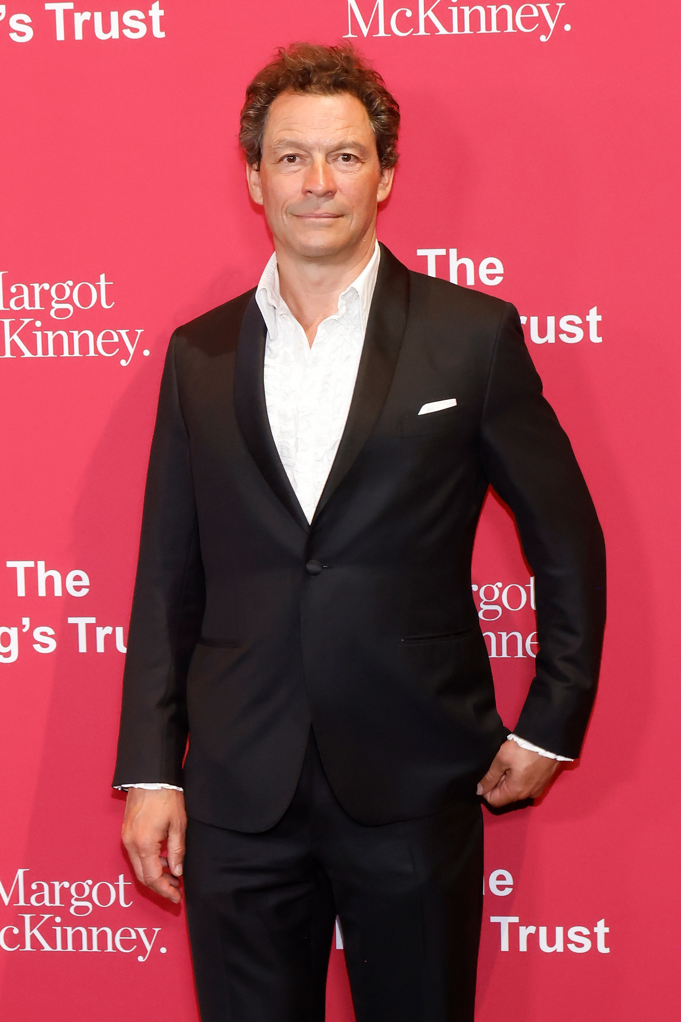 Dominic West in a fitted suit with a white shirt, posing with one hand in pocket
