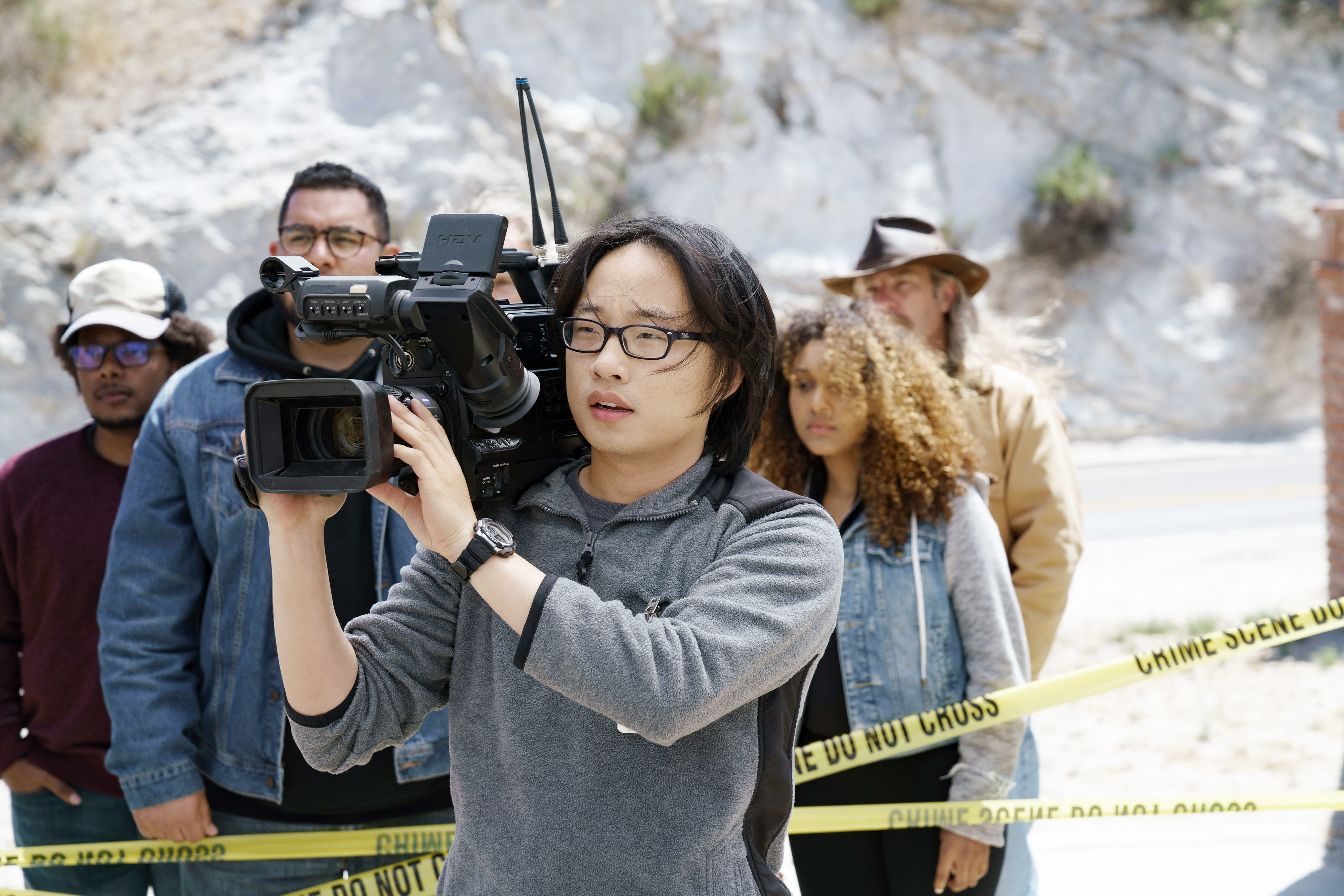 Jimmy O. Yang holding a video camera with others in the background behind yellow caution tape
