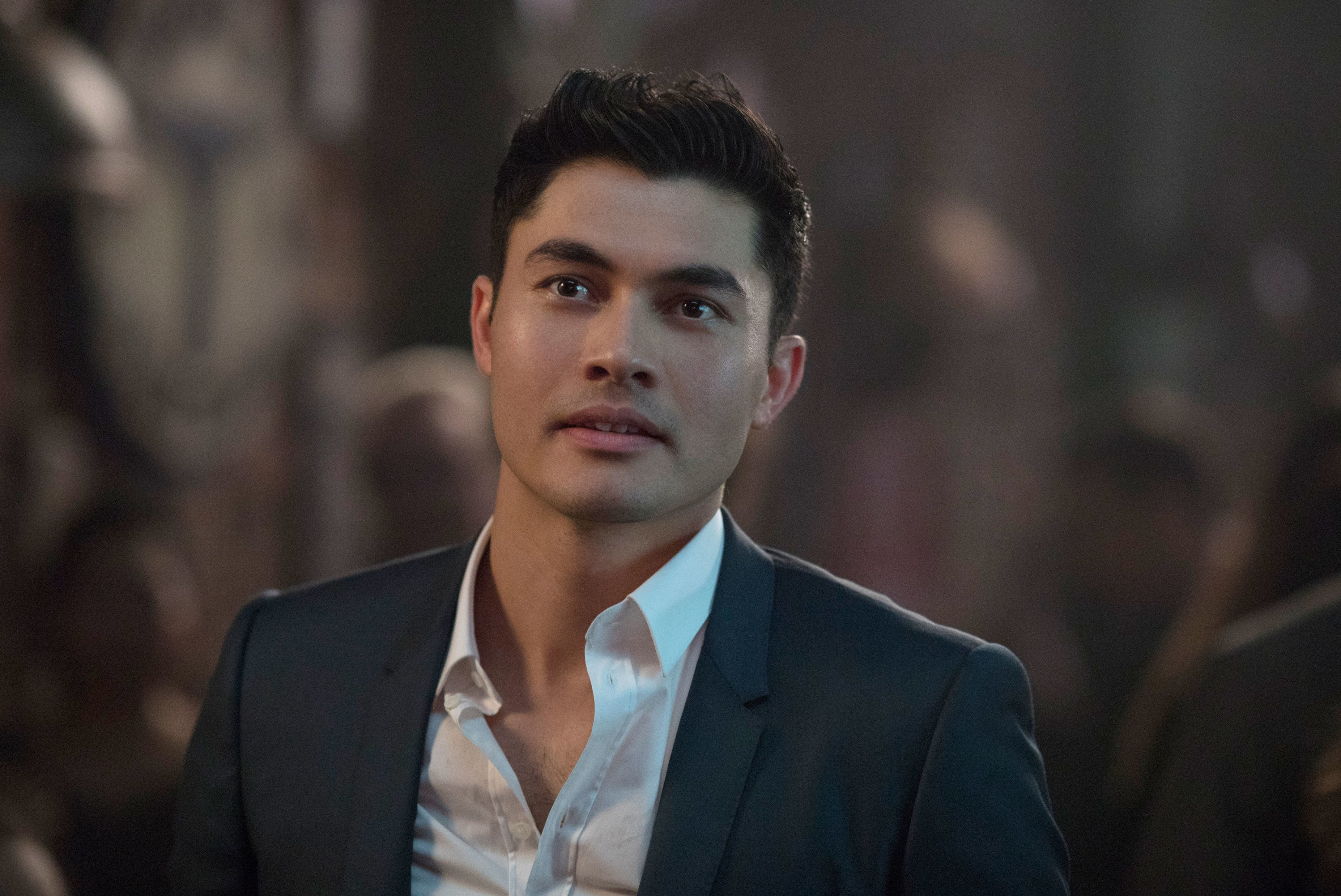 Henry Golding in a sharp suit looking pensive against a blurred background