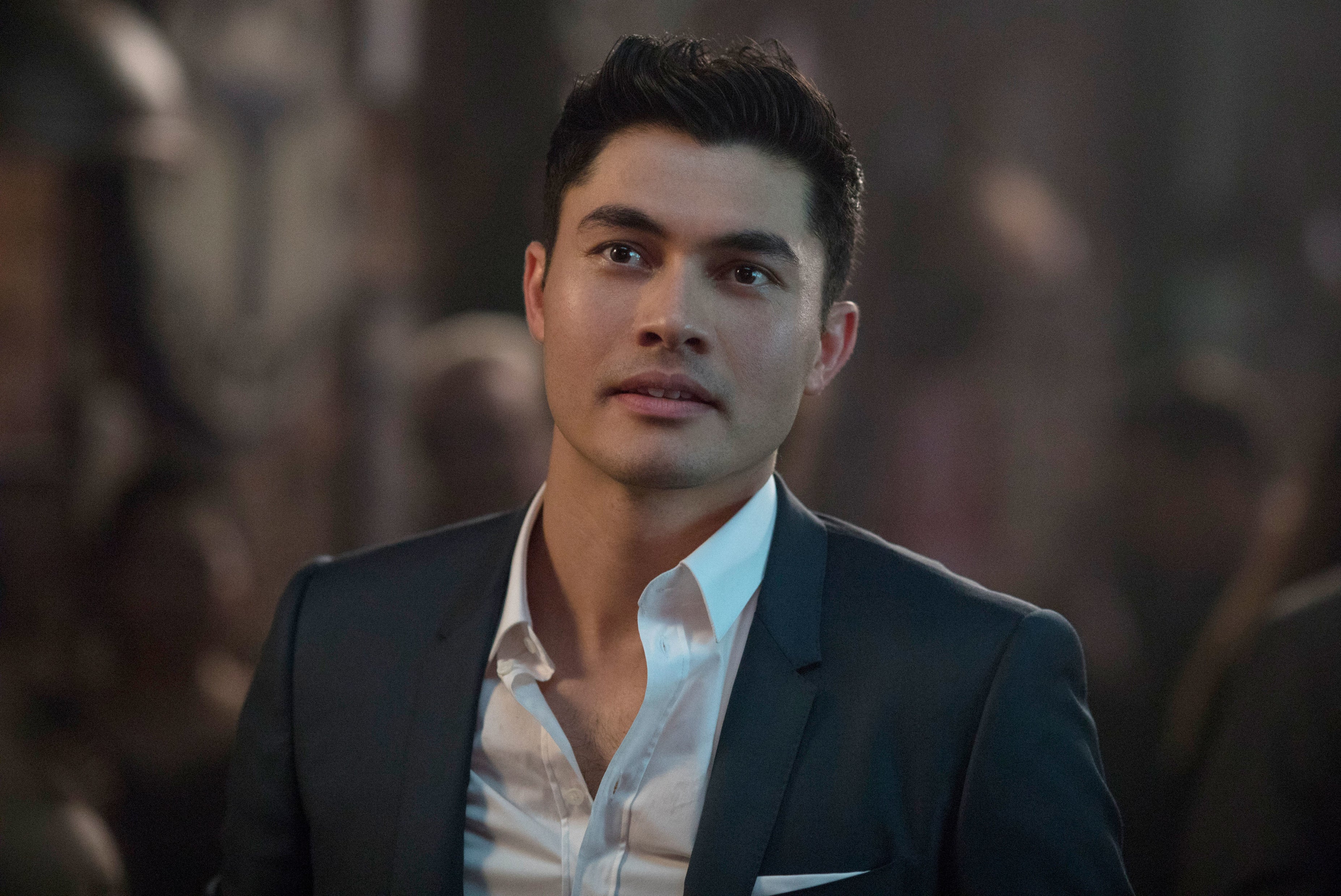 Henry Golding in a sharp suit looking pensive against a blurred background