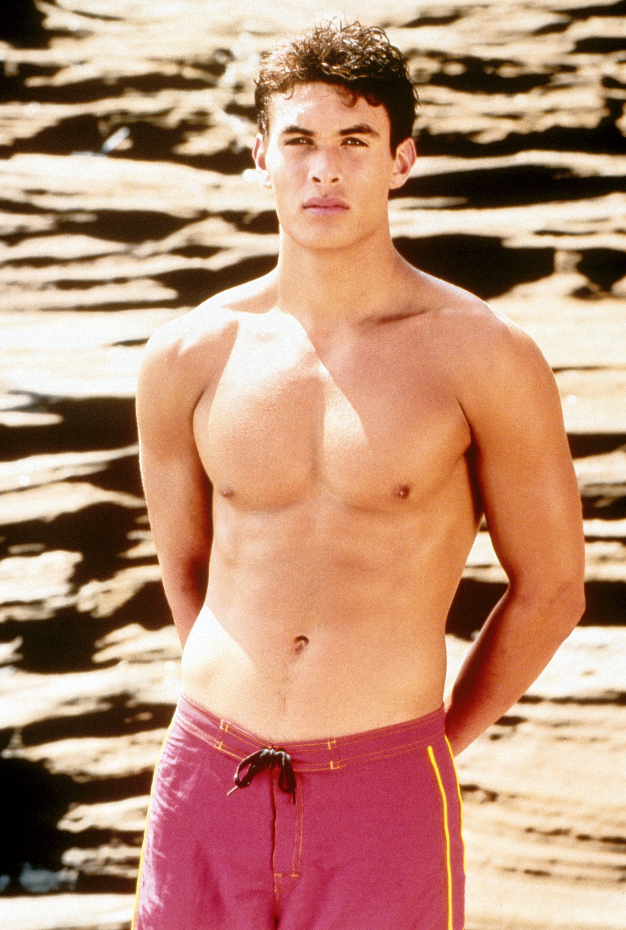 Jason Momoa shirtless on a beach wearing swim shorts