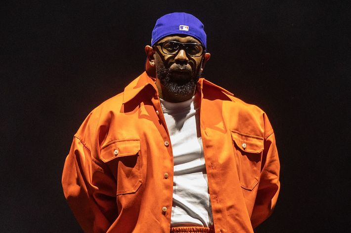 Man on stage wearing a blue cap and orange jacket, giving a performance