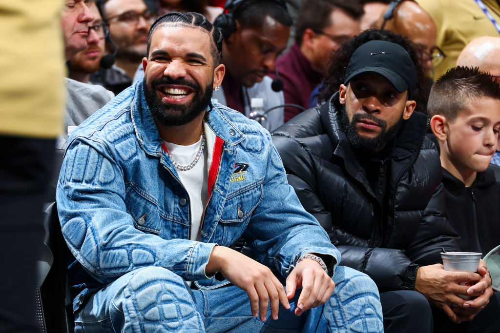 Drake wearing a denim jacket and jeans smiles while sitting courtside at a basketball game