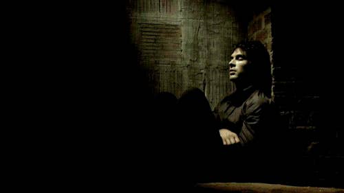 Damon sitting with knees drawn up, looking to the side, in a dimly lit room