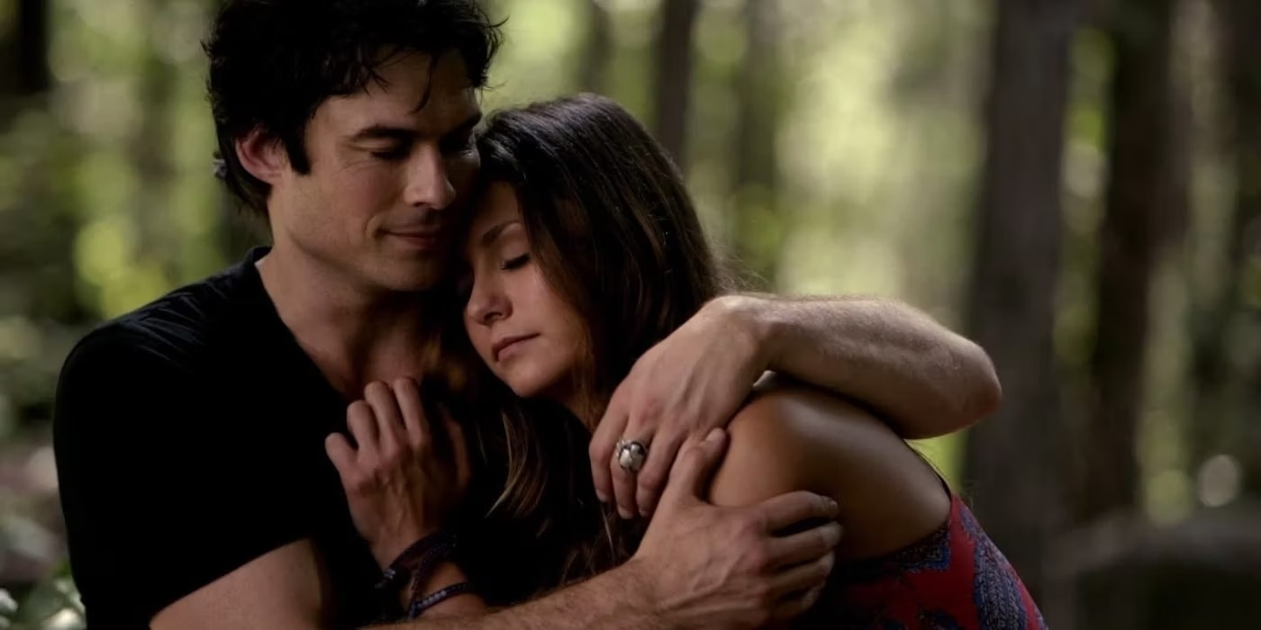 Two characters, Damon and Elena from the TV show &quot;The Vampire Diaries,&quot; are embracing each other in a forest setting