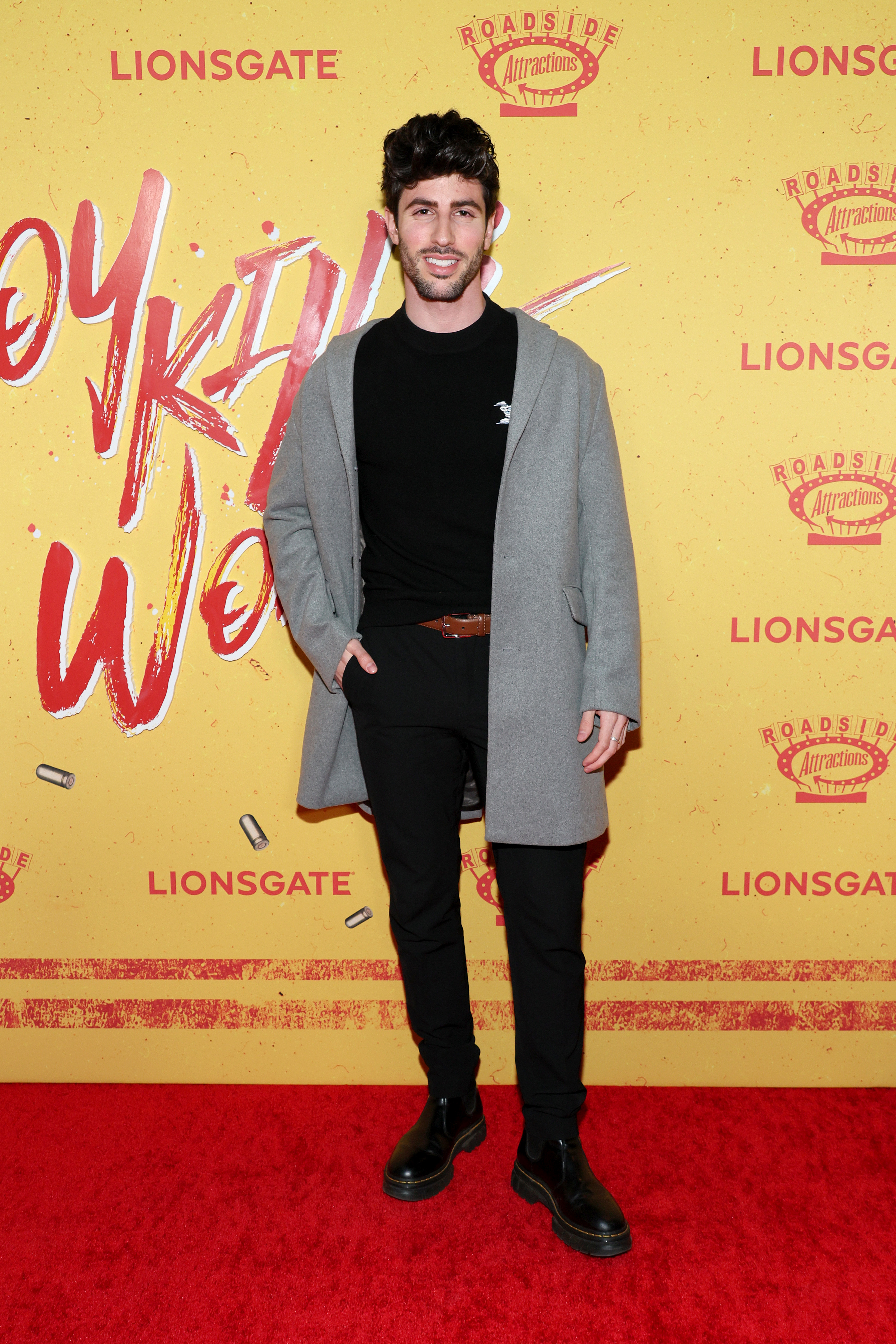 Man in black turtleneck, pants, and boots with gray coat posing on event backdrop