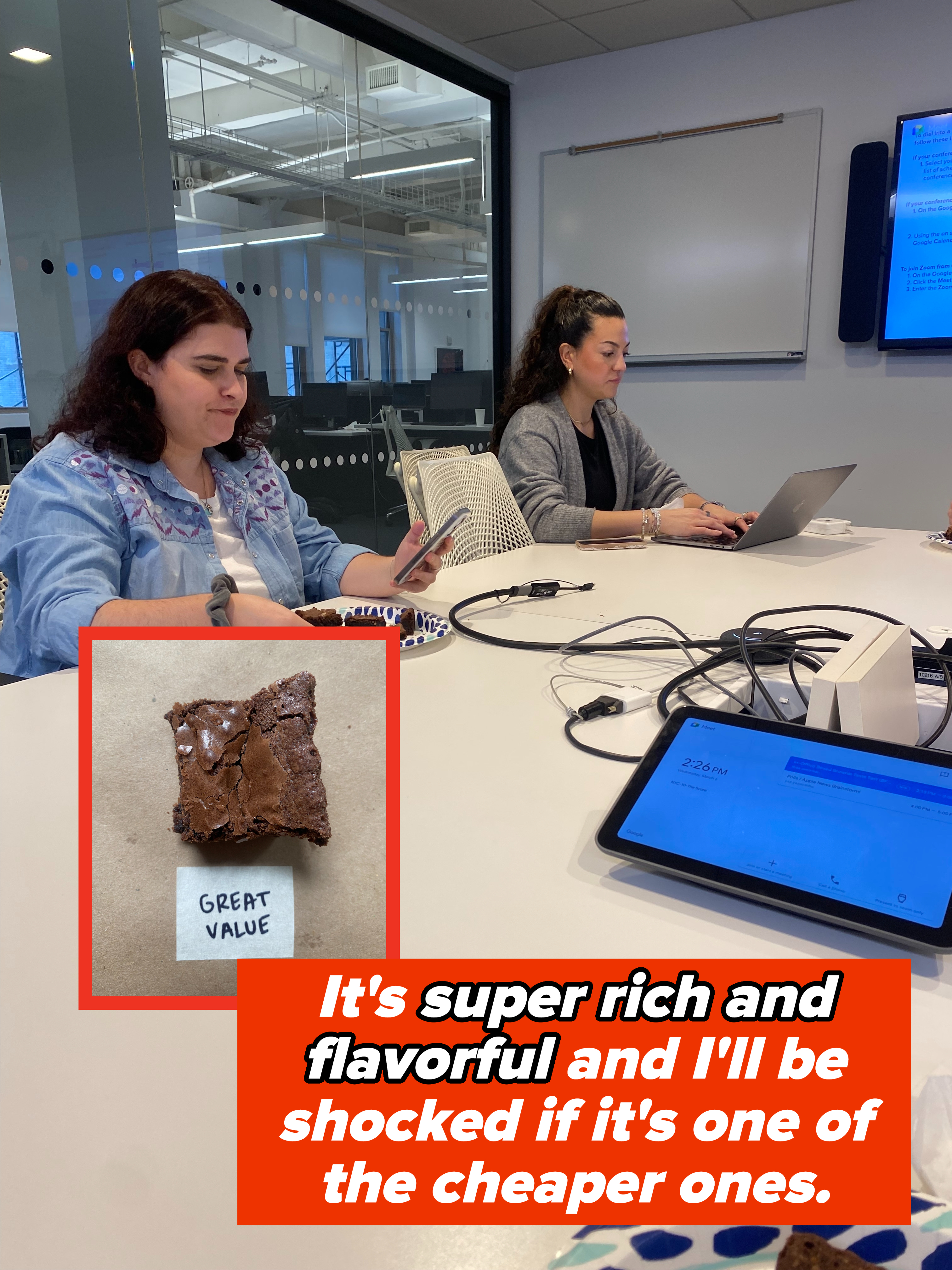 Two people at a table with laptops, one holding a plate of food, in a modern office setting