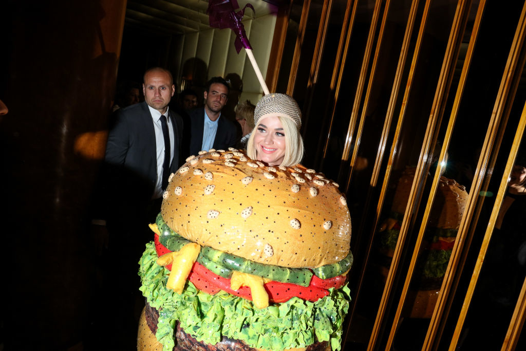 Katy in a hamburger costume with a sesame seed bun design and toppings; two individuals in the background
