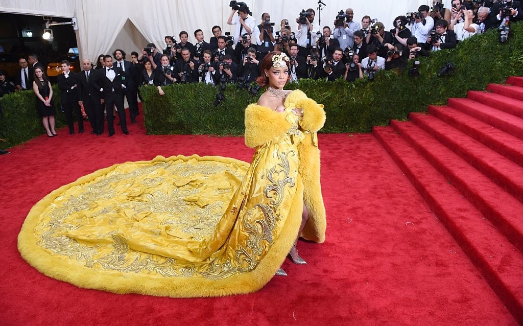 Rihanna in elaborate gown with long train posing on the red carpet, photographers in background