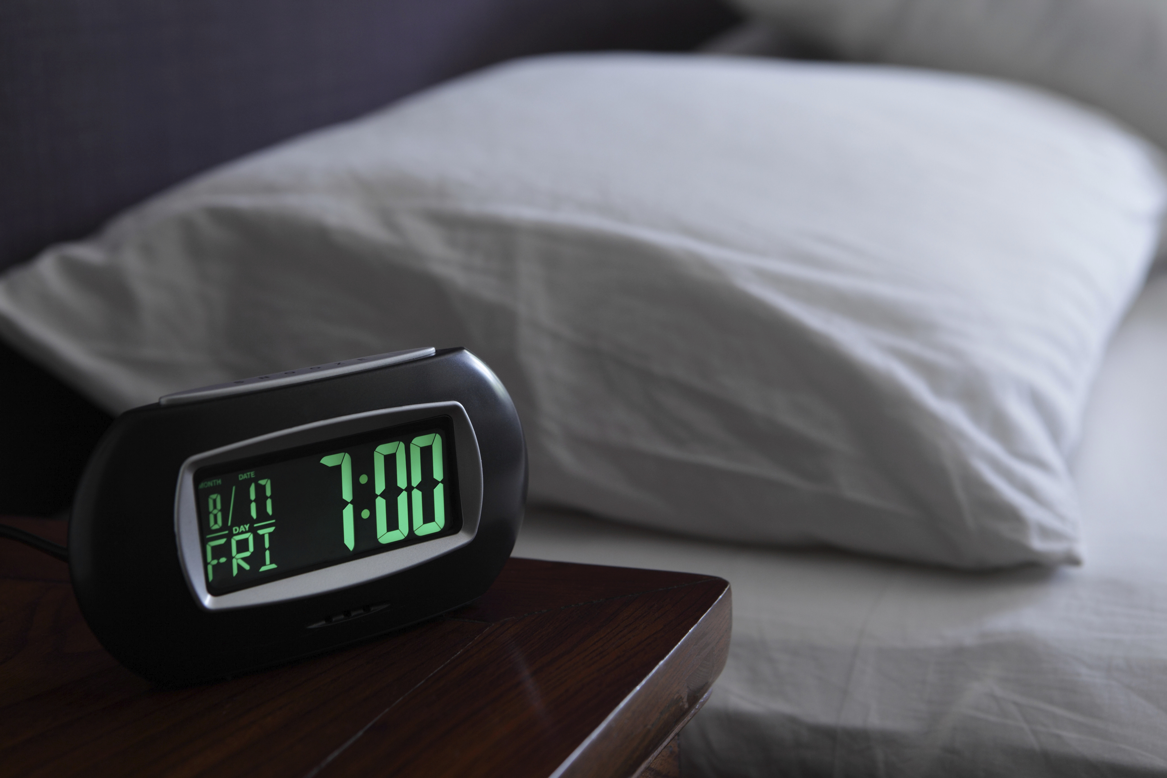 Digital alarm clock shows 7:00 AM next to a bed with white pillows